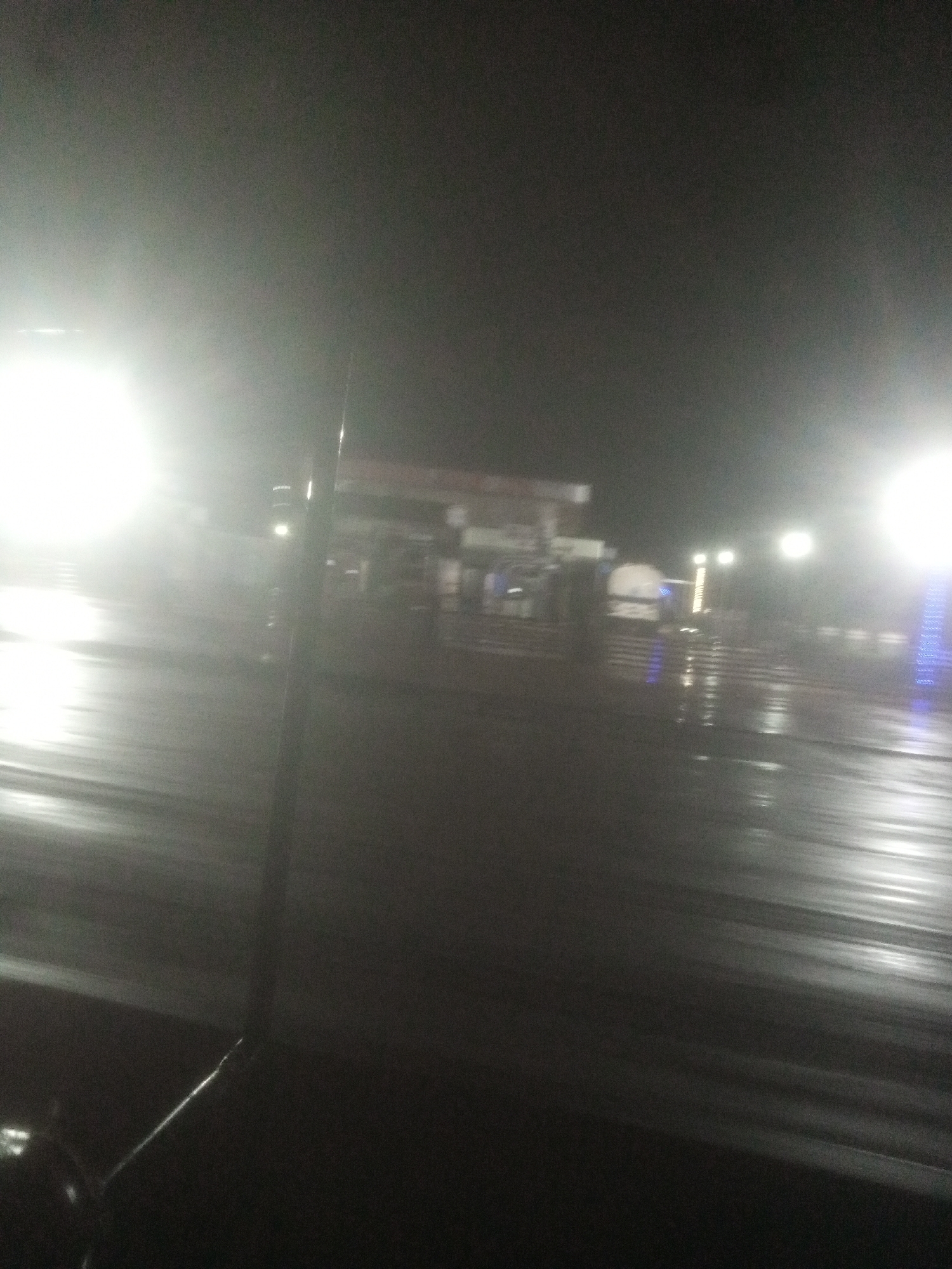 The image depicts a dimly lit, possibly rainy night scene at a gas station in Tundun Wada South, Nigeria. Bright lights from the gas station illuminate the wet pavement, creating reflections and a hazy atmosphere. The overall impression is one of low visibility and a nocturnal urban setting.