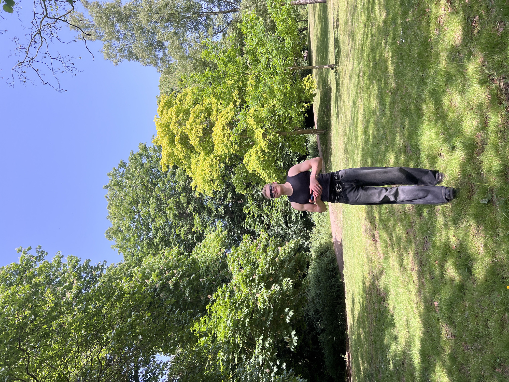 The image shows a person standing on a grassy area in Ipswich, United Kingdom. The person is wearing a black tank top, black pants, and sunglasses. They have their arms crossed. Behind the person are trees and a blue sky. The grassy area appears to be a park or garden.