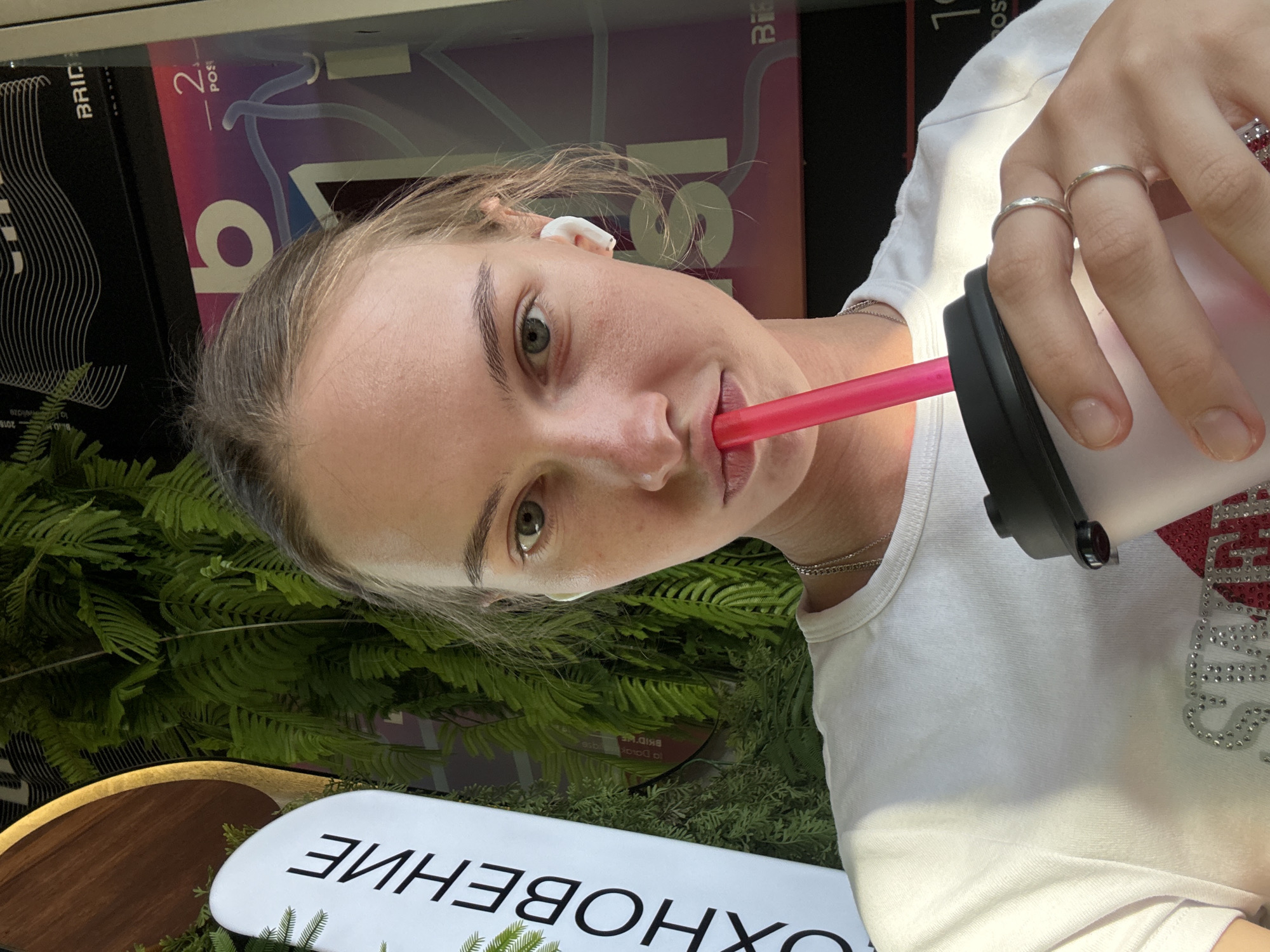 A young woman with fair skin, light brown hair, and light green eyes is shown in a close-up, selfie-style shot. She is wearing a white t-shirt, a silver necklace, and two silver rings on her right hand. She has white earbuds in both ears and is holding a pink straw to her mouth, with the straw extending into a clear cup with a black lid. The background is blurred but appears to be an outdoor setting with lush green foliage, possibly ferns, and signage with text in Russian. The signage includes the word "ВХОЧЕНИЕ" and other text that is partially obscured. The overall lighting suggests it is daytime. There is no identifiable information in the image that would indicate the specific location within the city of Perm, Russia.