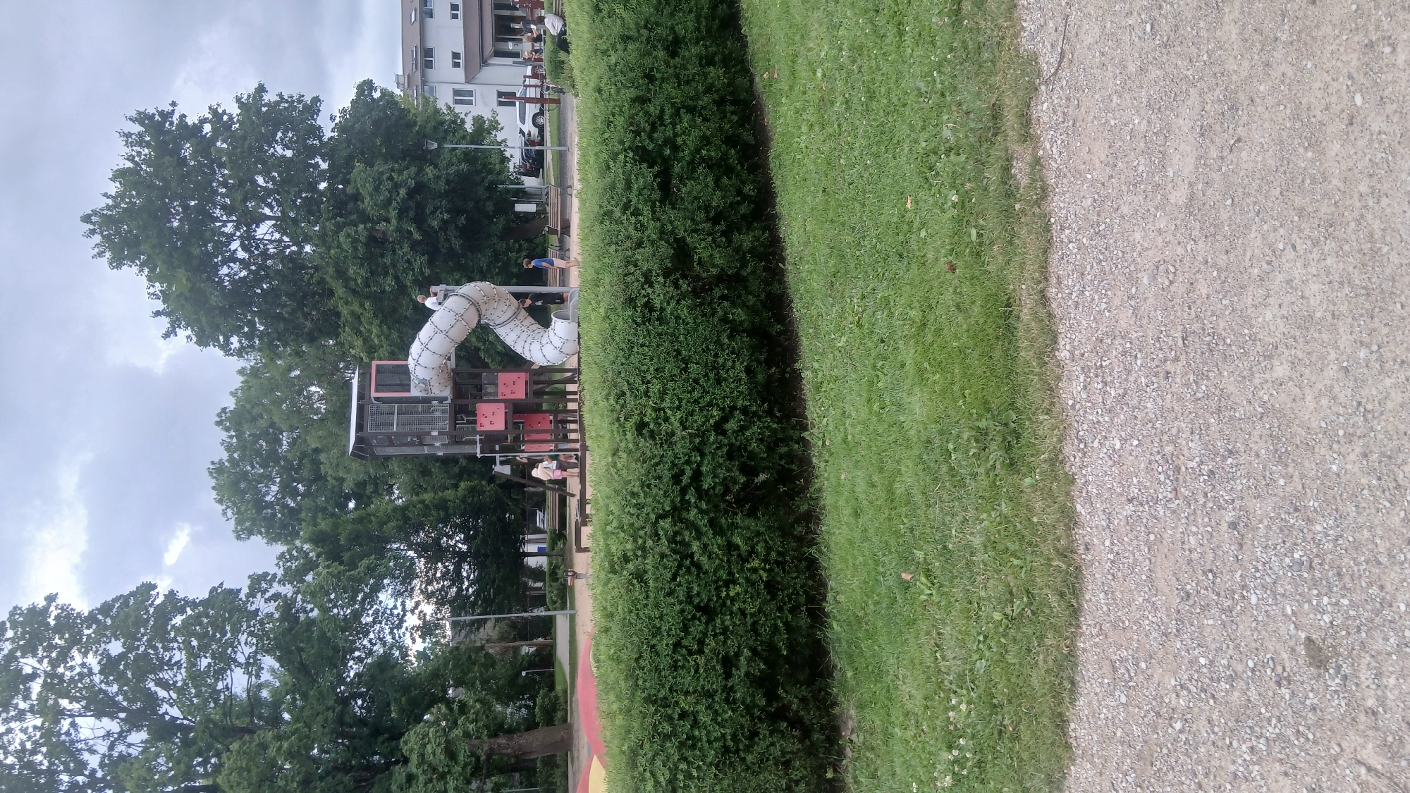 A playground with a modern, multi-level structure featuring a spiral tube slide is the focal point of this outdoor scene. The structure has a central tower with metal grating and pink accents. Several children are visible near the base of the structure, with one child appearing to be on the slide. A tall, dense green hedge runs across the foreground, separating the playground area from a grassy lawn and a gravel path. Trees, including a large, mature one on the left, provide shade and a natural backdrop. In the background, a white building with several windows and a few parked cars are visible, suggesting an urban or suburban setting. The sky is overcast with clouds. The overall impression is a well-maintained public park or playground. This scene is likely located in Jelgava, Latvia, given the context of the prompt.
