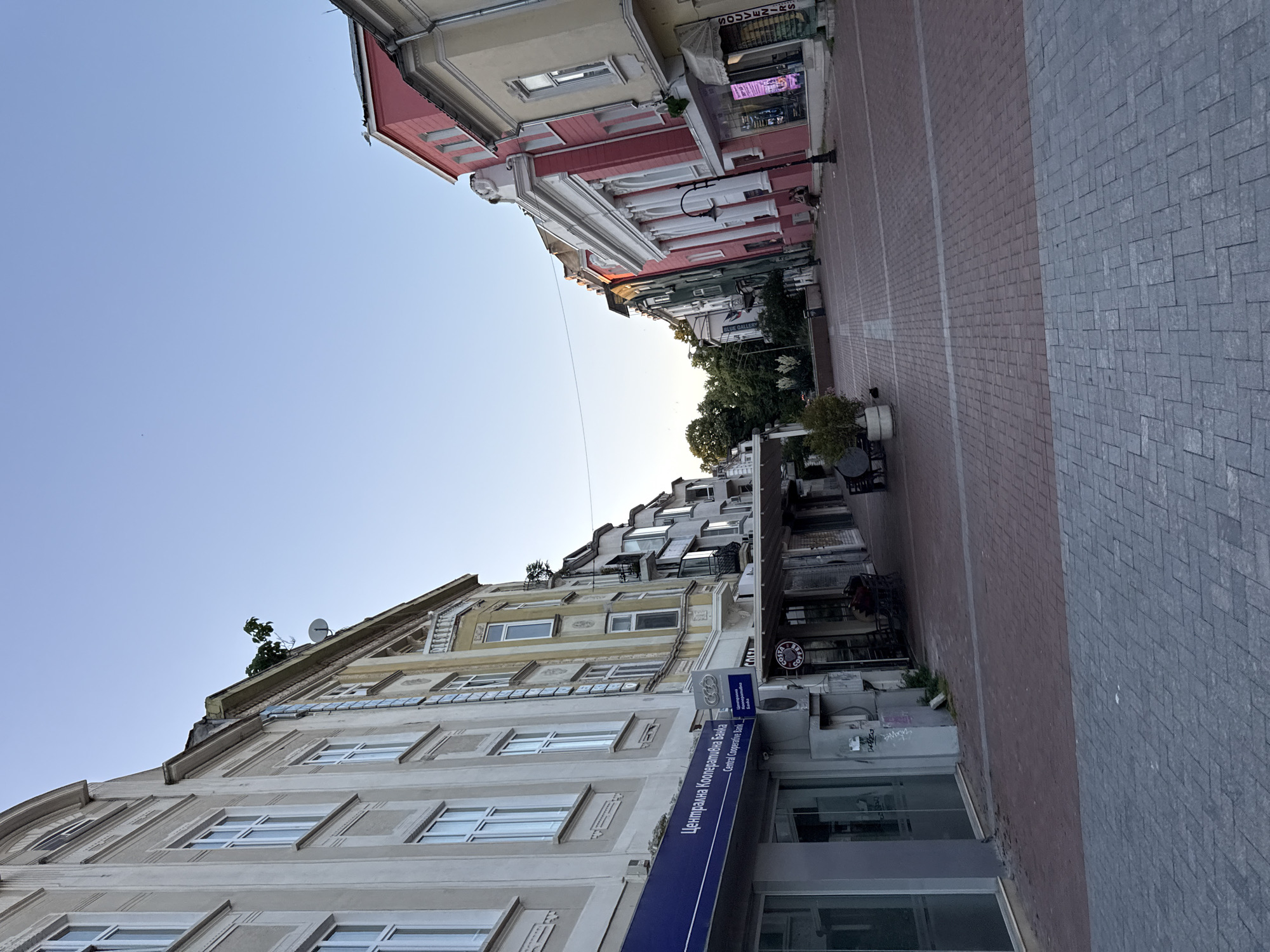 The image depicts a pedestrian street in an urban setting, likely in a European city, given the architecture and signage. The street is paved with brick and stone tiles. On the left, a building features a large blue sign with white text that reads "Централна Кооперативна Банка" and "Central Cooperative Bank." Adjacent to this are storefronts with large glass windows. Further down the street, on the right, a vibrant pink building with white classical columns stands out. Other buildings along the street are painted in various colors, including yellow and green, and feature balconies and multiple windows. Some shops have awnings. There are trees lining the street in the distance, and the sky is clear and blue. A few people are visible sitting on a bench in front of the pink building. The overall atmosphere suggests a quiet street, possibly in the early morning or late afternoon, with minimal activity.