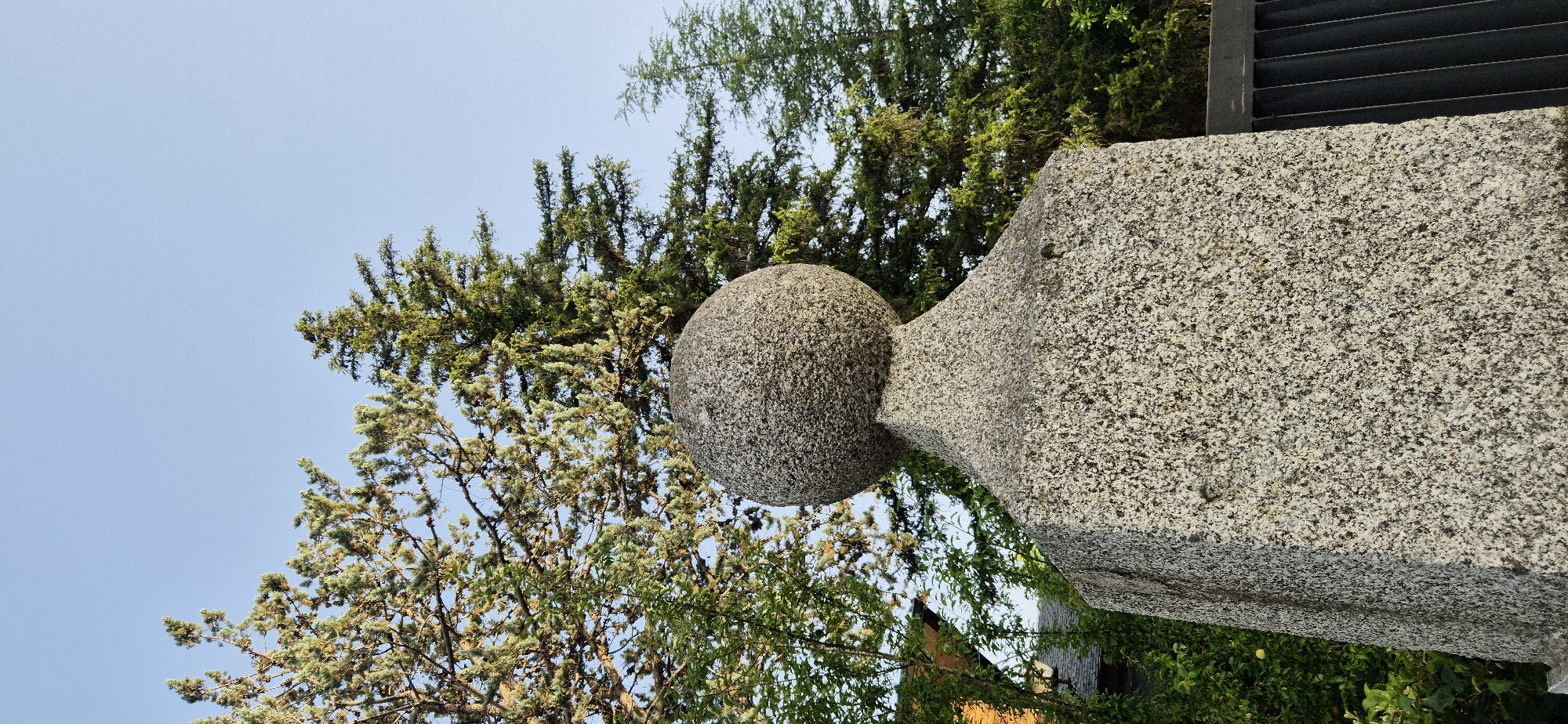 A close-up, low-angle shot captures the top portion of a granite post capped with a granite sphere, against a clear blue sky. The post is square and textured, tapering to a point where the sphere is affixed. The sphere is also textured and appears to be carved from a single piece of granite. Behind the post, lush green trees and foliage form a dense backdrop, with branches extending into the sky. Part of a building, with an orange facade, is visible to the left, peeking through the greenery. To the right of the post, a black metal fence with vertical slats is partially visible. The lighting suggests it is daytime, with soft illumination on the granite and the foliage.