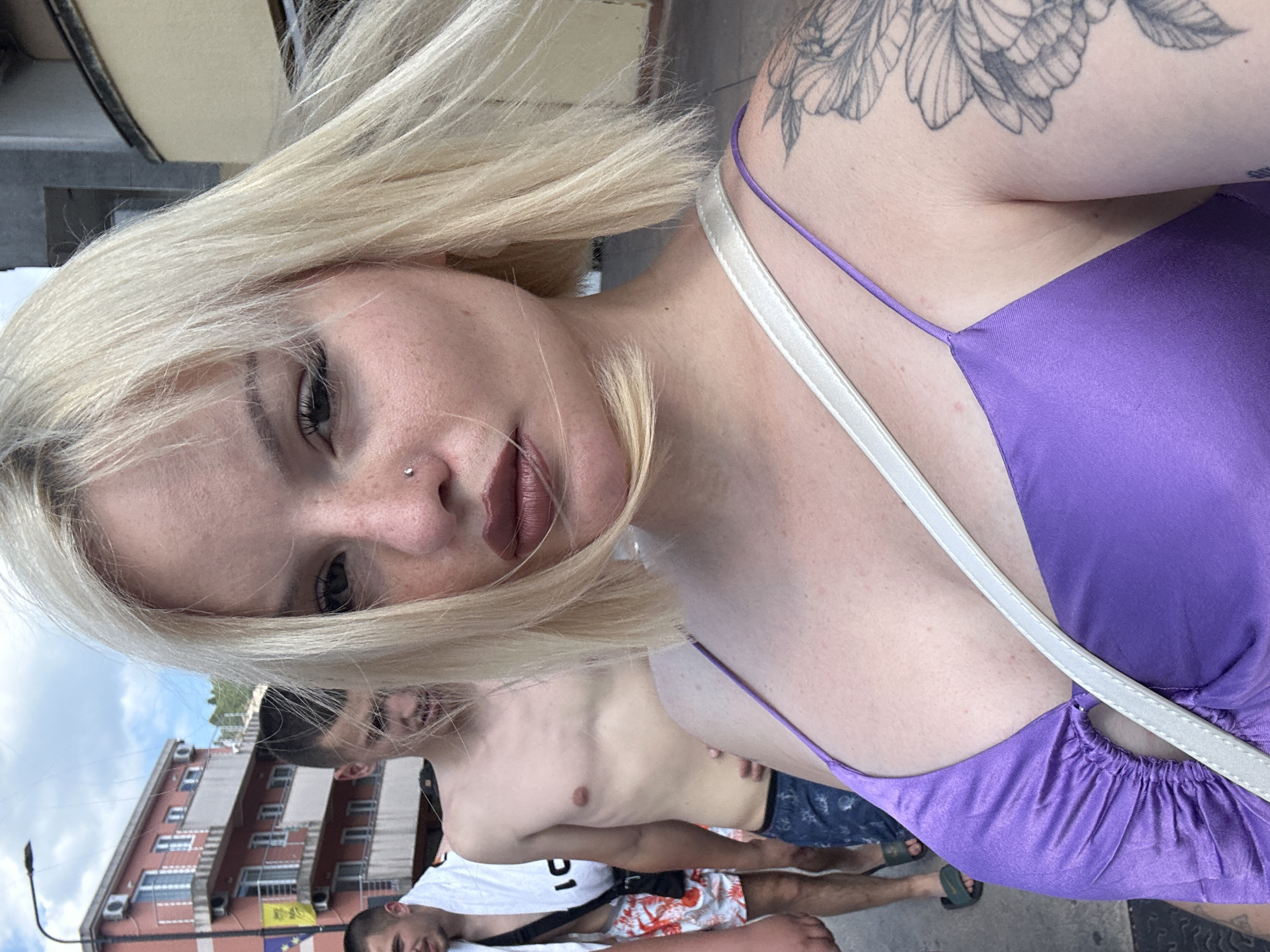 A group of people are walking on a sidewalk in front of a building. The person in the foreground is a woman with blonde hair and freckles, wearing a purple dress. She is looking at the camera and taking a selfie. Behind her, two men are walking. One man is wearing a white towel over his shoulder and floral shorts, and the other man is shirtless and wearing blue shorts. The building behind them appears to be a hotel with balconies. The scene is set in Budva, Montenegro.