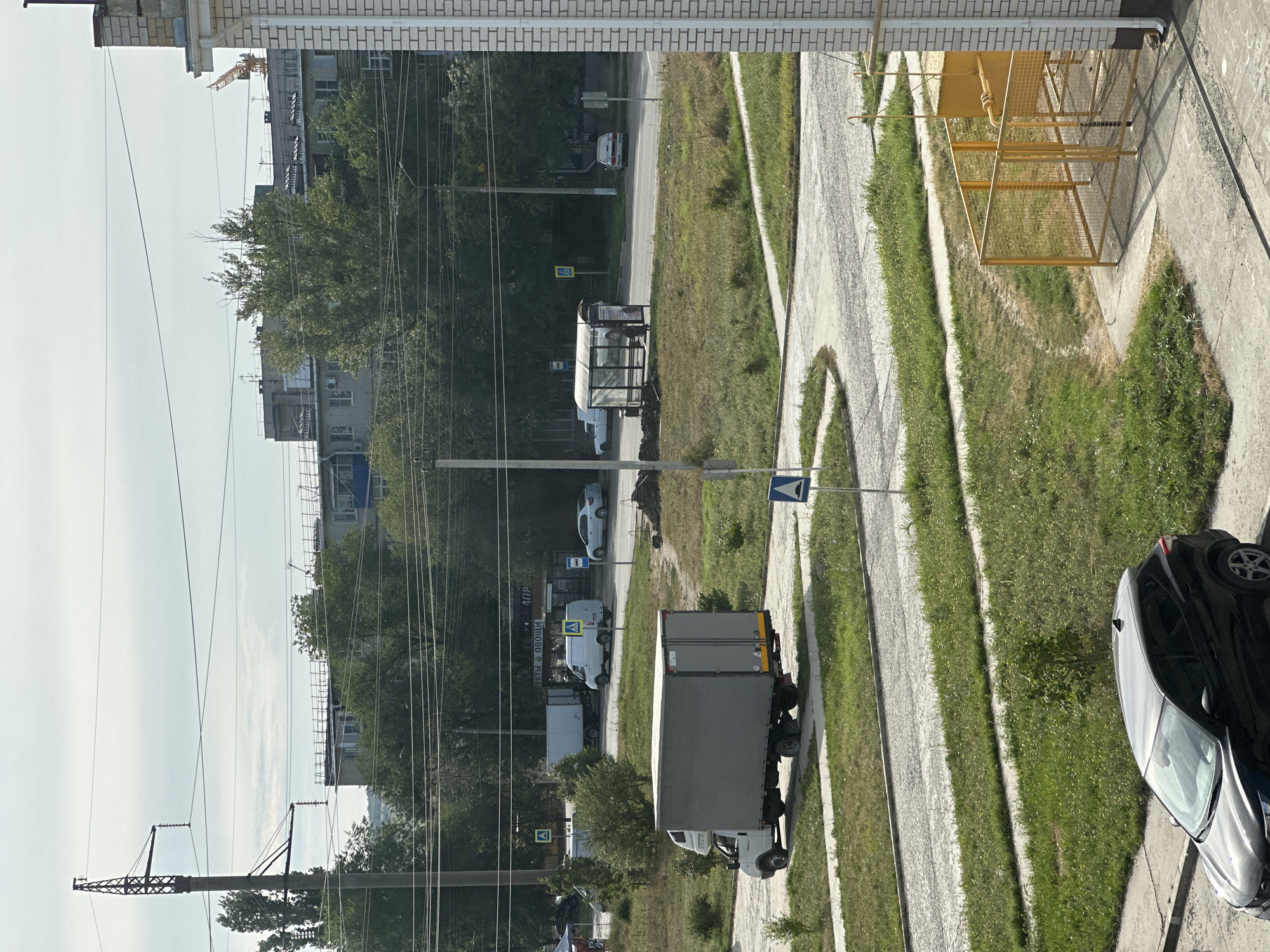 The image captures a street scene in Nevinnomyssk, Russia, under an overcast sky. In the foreground, a black SUV is parked on a concrete surface. To the right, a yellow industrial tank is enclosed by a metal fence, adjacent to a brick building.
The midground features a paved road with a bus stop shelter and a traffic sign. Several vehicles are present, including a white box truck with a grey cargo area, two white vans, and two white cars. A few trees and green grassy areas are visible. In the background, a cluster of apartment buildings and a construction crane are discernible, along with power lines and a tall electricity pylon. The overall environment appears to be a residential and commercial area.