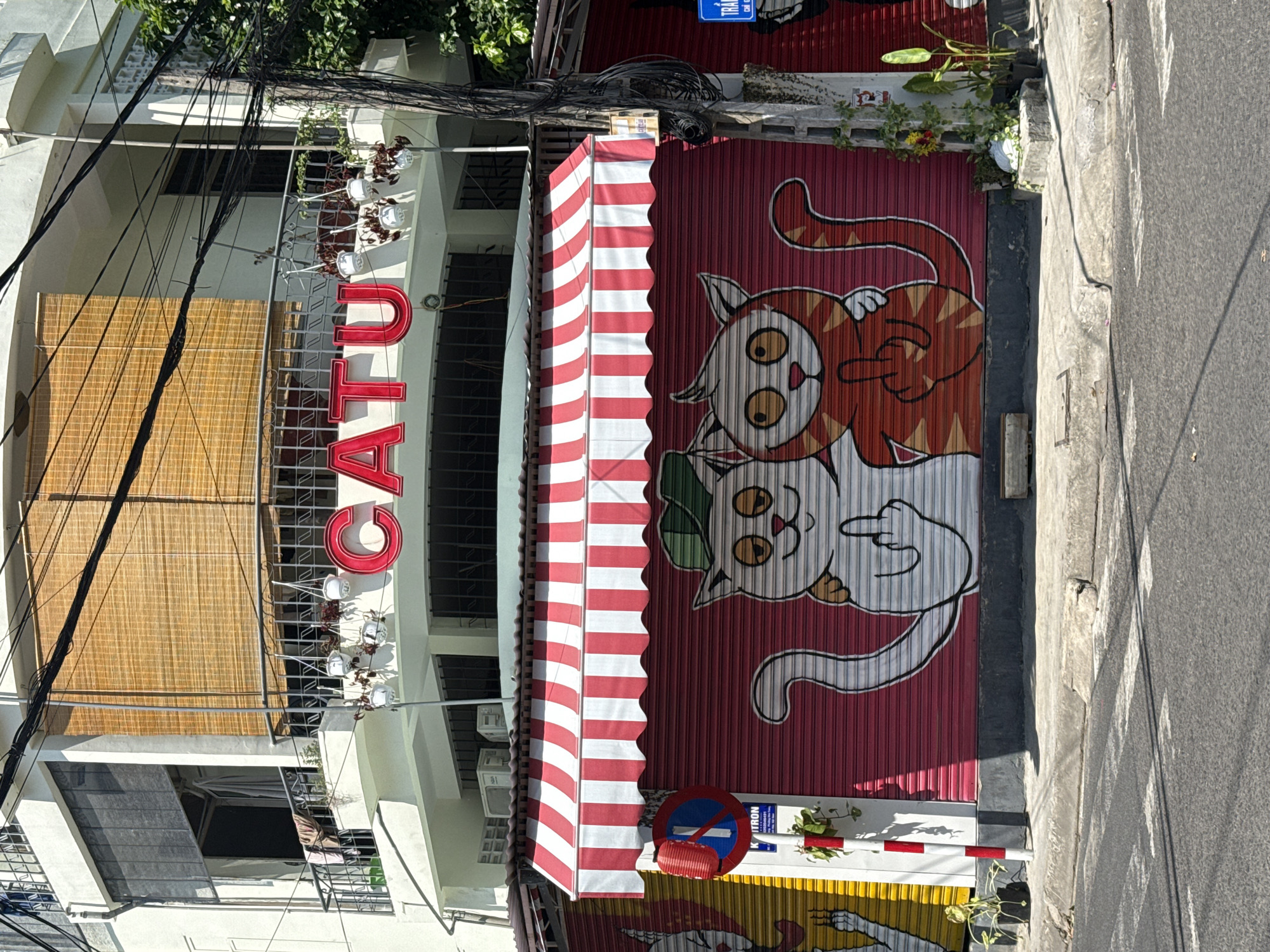 The image shows the storefront of a business named "CATU" in Nha Trang Ward, Vietnam. The building has multiple stories, with a white facade and a large, red neon sign spelling "CATU" above the ground floor. The ground floor features a red and white striped awning covering a closed metal roll-up door. This door is decorated with a mural of two cartoon cats, both giving the middle finger. One cat is white with a green hat, and the other is orange with tiger stripes. In front of the roll-up door, there is a traffic sign and a red and white striped pole with a potted plant. The street in front of the building is paved and appears to be a city street. The overall scene suggests a vibrant and potentially quirky establishment.