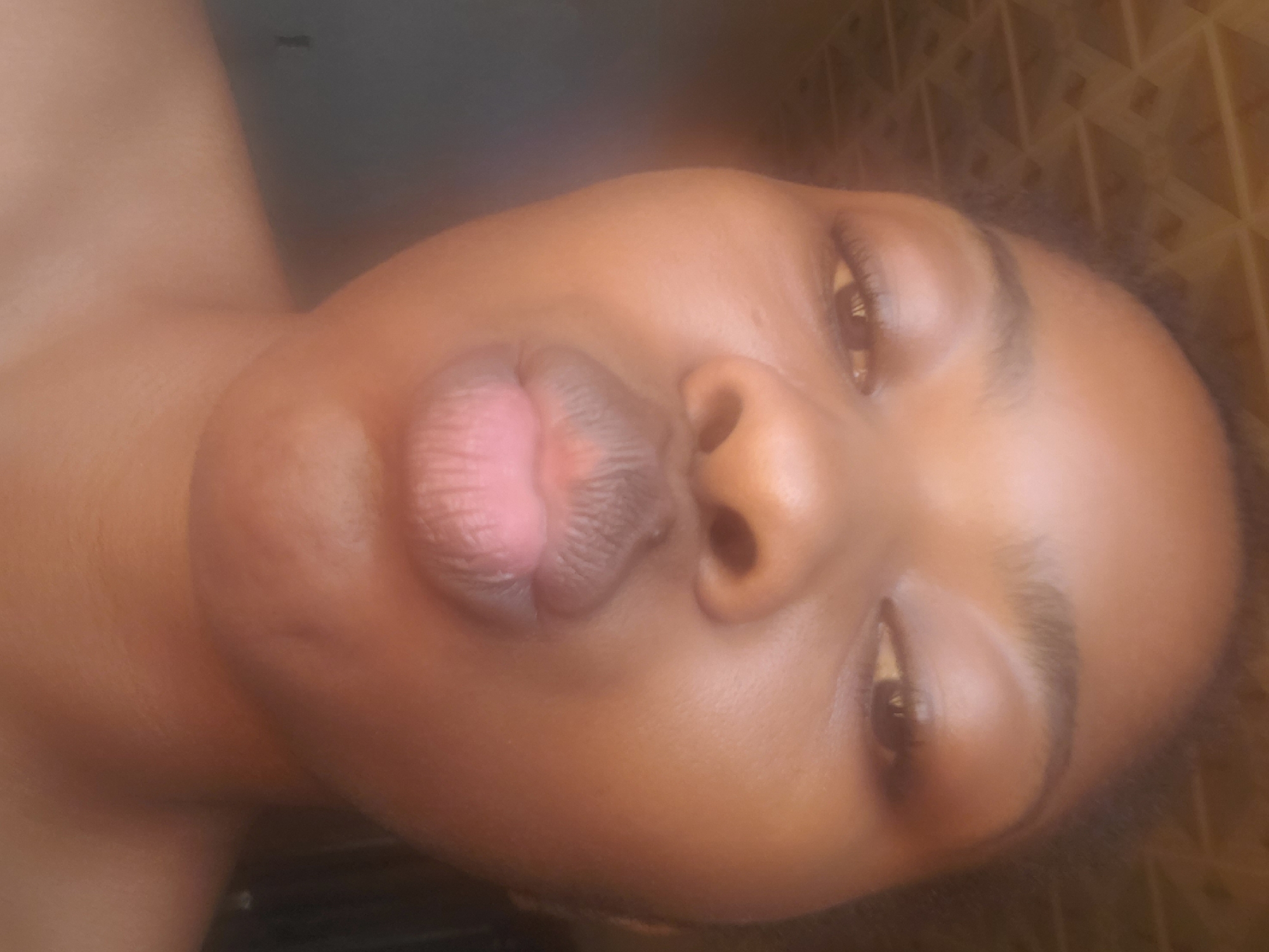A close-up portrait features an individual, likely female, with a dark complexion, facing the camera directly. The subject has dark, textured hair visible around the top and sides of the head. Their dark eyes are open, with visible pupils and eyelashes. The eyebrows are well-defined. The subject's lips are significantly pursed into a "kissing" or "duck face" pose. No specific clothing or accessories are visible, only bare skin around the neck and shoulders.
The setting appears to be indoors. In the upper left background, a light-colored, patterned surface is visible, exhibiting a repeating geometric (possibly triangular or square grid) texture, which could be a ceiling or wall. The remainder of the background is indistinct, appearing darker and out of focus, suggesting a shadowed interior or plain dark surface. Lighting appears to be soft and frontal, illuminating the subject's face. The image does not contain any visual markers or details to confirm a specific geographical location such as Kakau, Nigeria.