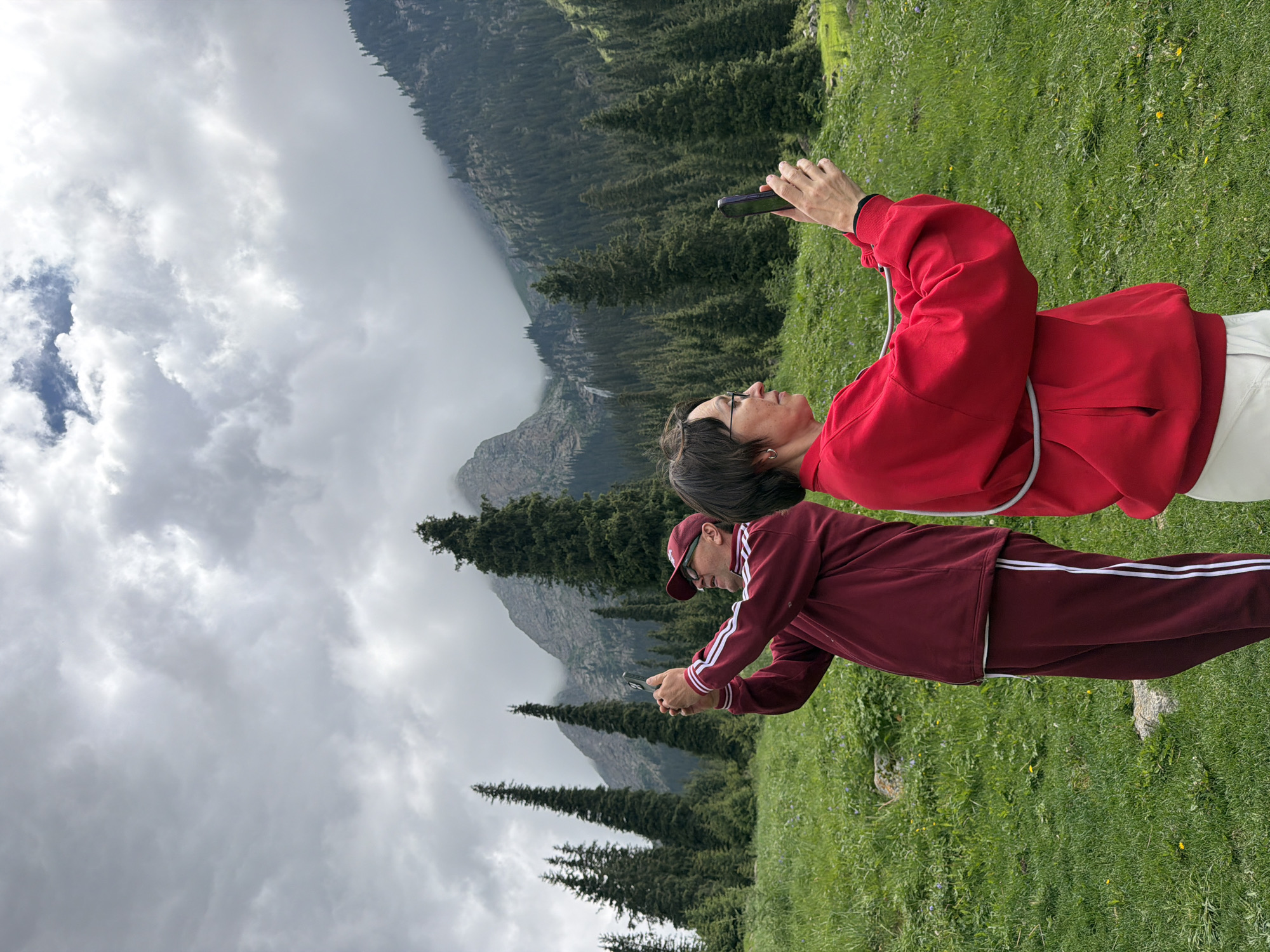 Two people are taking pictures of a mountain landscape with their phones. The person in the foreground, wearing a red sweatshirt and white pants, is holding a dark-colored phone horizontally. They are looking at the phone with a neutral expression and are positioned on a grassy hillside. Behind them, another person, wearing a maroon tracksuit and a baseball cap, is also looking at their phone, which they are holding in a similar fashion.
The background of the image shows a steep mountain slope covered with dense evergreen trees. Further in the distance, large, rocky mountains are visible, partially obscured by clouds. The sky is overcast with heavy gray clouds, suggesting a cool or possibly rainy day. The grass in the foreground is green and appears to be a meadow with a few small yellow flowers. The overall scene evokes a sense of exploration and appreciation for nature. No visible text is present in the image.