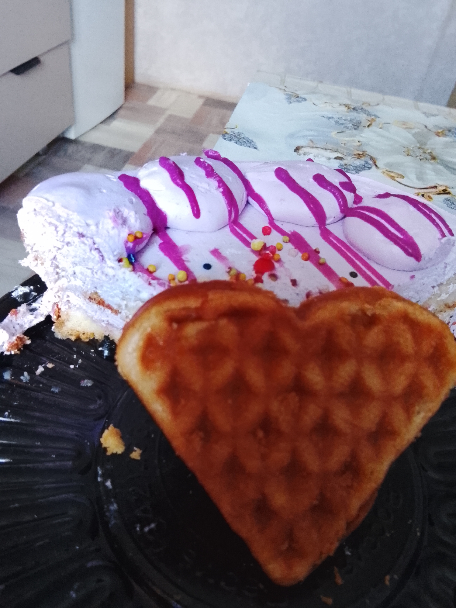 A heart-shaped waffle sits in the foreground on a black plastic plate, partially obscuring a slice of cake in the background. The cake is decorated with light purple frosting and a series of white dollops with purple stripes. Multicolored sprinkles are scattered on the cake. A white dresser is visible in the upper left corner, and a patterned tablecloth is behind the plate. The scene appears to be indoors. This is likely a residence in Kuyeda, Russia.
