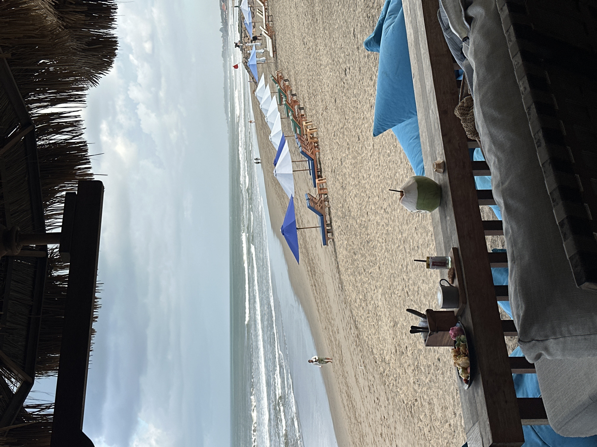 The image depicts a tranquil beach scene in North Kuta, Indonesia, likely during the late afternoon given the soft lighting. In the foreground, a wooden railing supports a small selection of items: a plate of colorful pastries, a cup of coffee, a glass with a straw, a small coaster, and a holder with utensils. Beside these, a young coconut with a straw sits on the railing. Behind the railing, large, plush blue cushions are visible, offering comfortable seating.
The middle ground of the image showcases a sandy beach lined with numerous lounge chairs and umbrellas. The umbrellas are a mix of white and blue, providing shade for the unoccupied chairs. A single person is seen walking along the shoreline in the distance.
The background features the ocean with gentle waves rolling onto the shore. The sky is overcast with a soft, diffused light, suggesting a peaceful atmosphere. The overall impression is one of relaxation and leisure, typical of a beachfront resort in Bali.