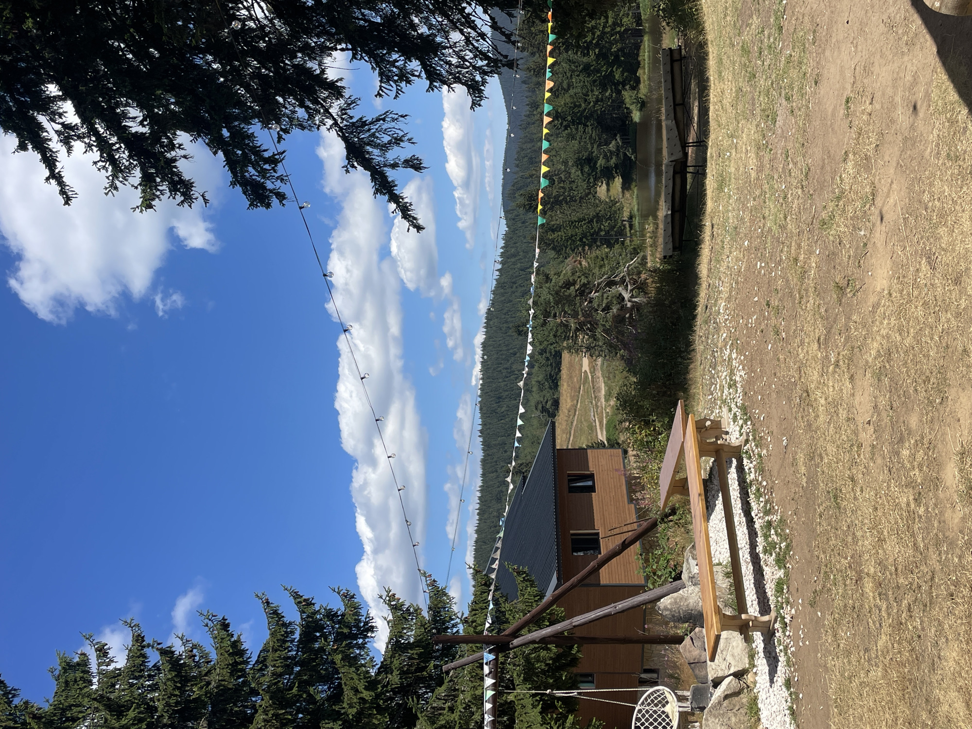This image captures a scenic outdoor setting likely near Smolyan, Bulgaria, on a bright, sunny day with a clear blue sky dotted with fluffy white clouds. In the foreground, dry, matted grass covers the ground, with a dirt path visible. A wooden picnic bench with a lighter colored top sits on a bed of white gravel to the left of the frame. Behind it, a rustic wooden house with a dark roof and black-trimmed windows is visible. Further back, a tranquil lake or pond is surrounded by dense evergreen forest that stretches across rolling hills. A string of decorative lights hangs across the scene, adding a festive touch. Another wooden bench is situated closer to the water. The scene suggests a peaceful, natural retreat.