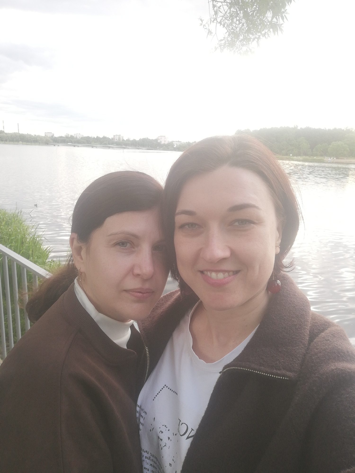 Two women are taking a selfie by a lake. The woman on the left has dark brown hair pulled back and is wearing a white turtleneck and a brown jacket. She is looking towards the camera with a neutral expression. The woman on the right has shorter brown hair and is wearing a white t-shirt with some text on it and a brown jacket. She is smiling at the camera. Both women appear to be in their 30s or 40s.
The background shows a body of water, with some trees and buildings visible on the opposite shore. There is also a railing in the foreground, suggesting the women are on a walkway or bridge. The sky is bright, with some clouds, indicating it might be daytime, possibly late afternoon, given the diffused sunlight. The overall scene is casual and relaxed. There is some text visible on the white t-shirt of the woman on the right, but it is not entirely legible. The location context is Kolpino, Russia.