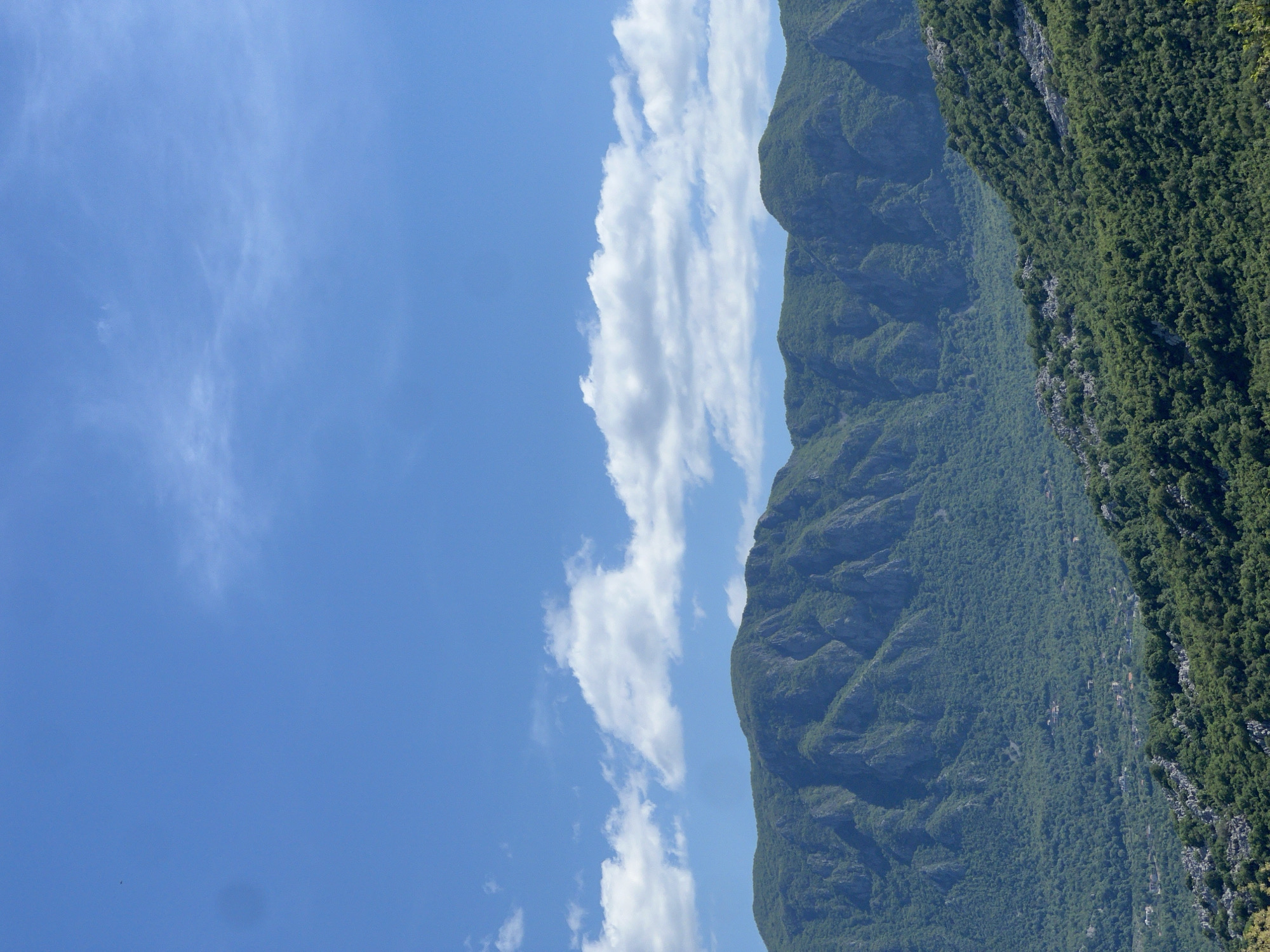The image depicts a landscape in Sotonići, Montenegro. Dominating the right side are steep, green mountains covered in dense trees. To the left of the mountains is a blue sky with scattered white clouds.