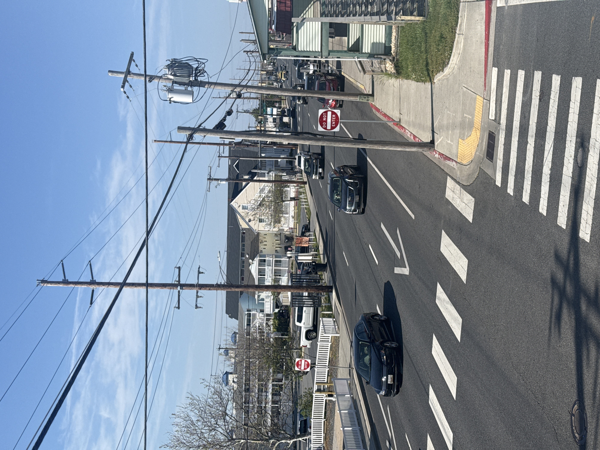 The image depicts a street scene in Ocean City, United States of America. It shows a street with asphalt pavement, marked with white crosswalk lines and directional arrows. Vehicles, including cars and a white van, are visible along the street. On either side of the street, there are sidewalks, utility poles with electrical wires, and buildings. Street signs, including "DO NOT ENTER" signs, are present. The sky is clear and blue. The scene appears to be during daytime, with shadows cast by the objects.