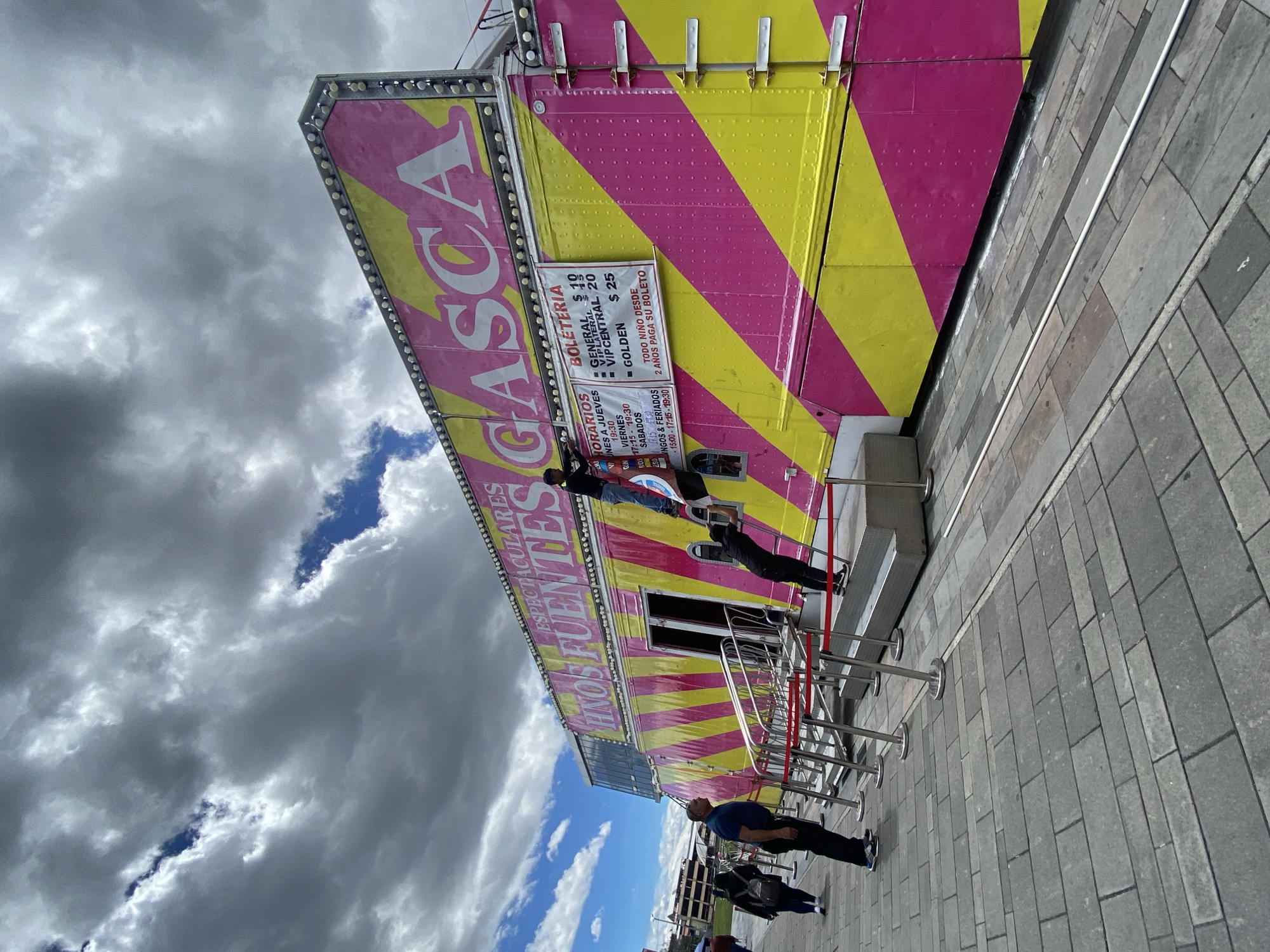The image shows a view of a "Hnos Fuentes Gasca" carnival attraction, recognizable by its bright pink and yellow striped design. The carnival structure displays ticket information and show times in Spanish. A queue of people is waiting outside the attraction, separated by metal barriers with red ropes. Several people are visible: some are standing in line, while others are walking past. The ground is paved with gray bricks. The sky is cloudy, with patches of blue visible.