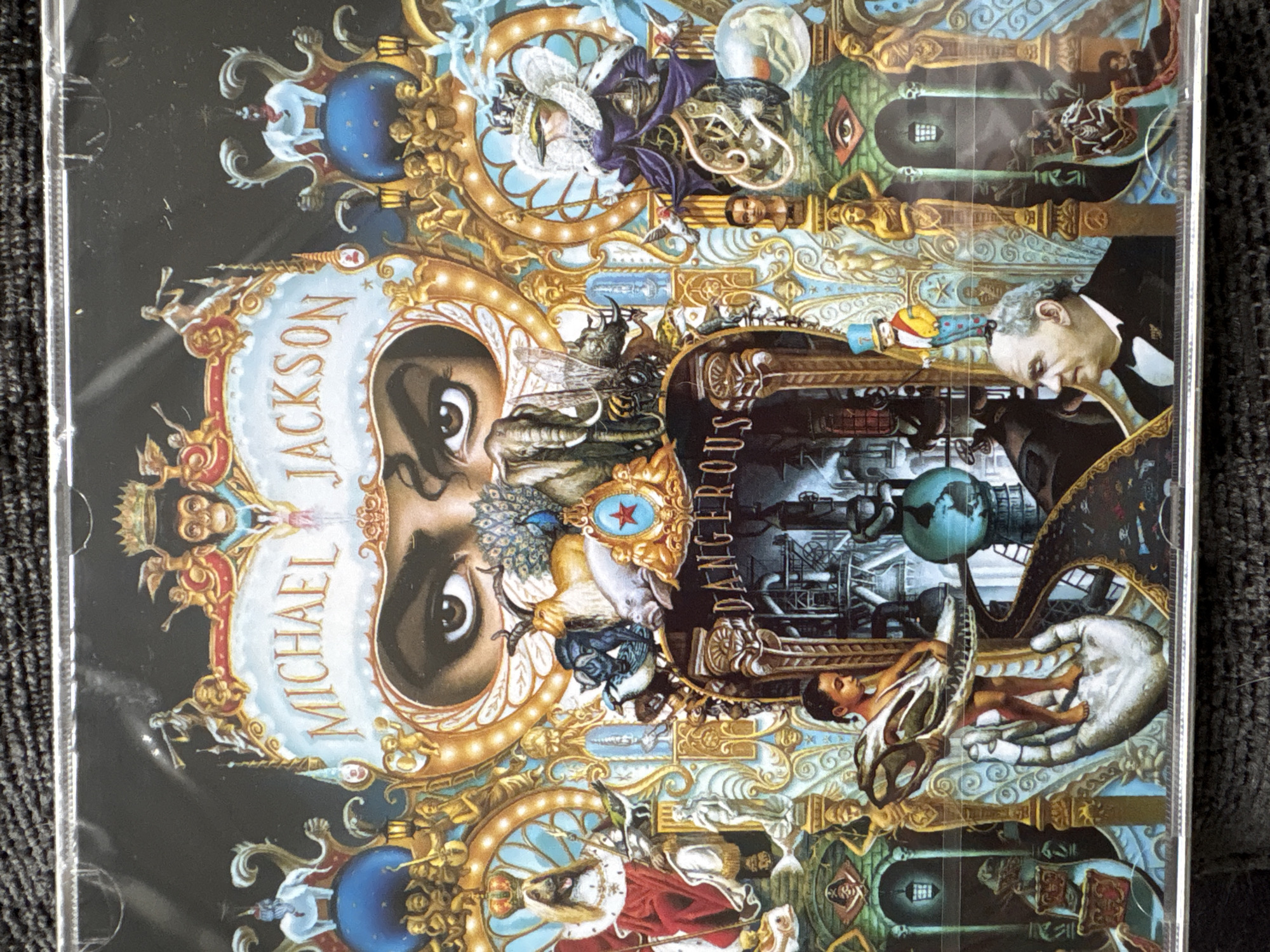 The image shows the cover art for Michael Jackson's album "Dangerous," rotated 90 degrees clockwise. The artwork is complex and fantastical, featuring Michael Jackson's face integrated into a collage of various symbols, figures, and scenes. The words "MICHAEL JACKSON" and "DANGEROUS" are visible within the design. The artwork is predominantly gold, black, and blue. The image does not show any scene located in the city of Manchester, United Kingdom.