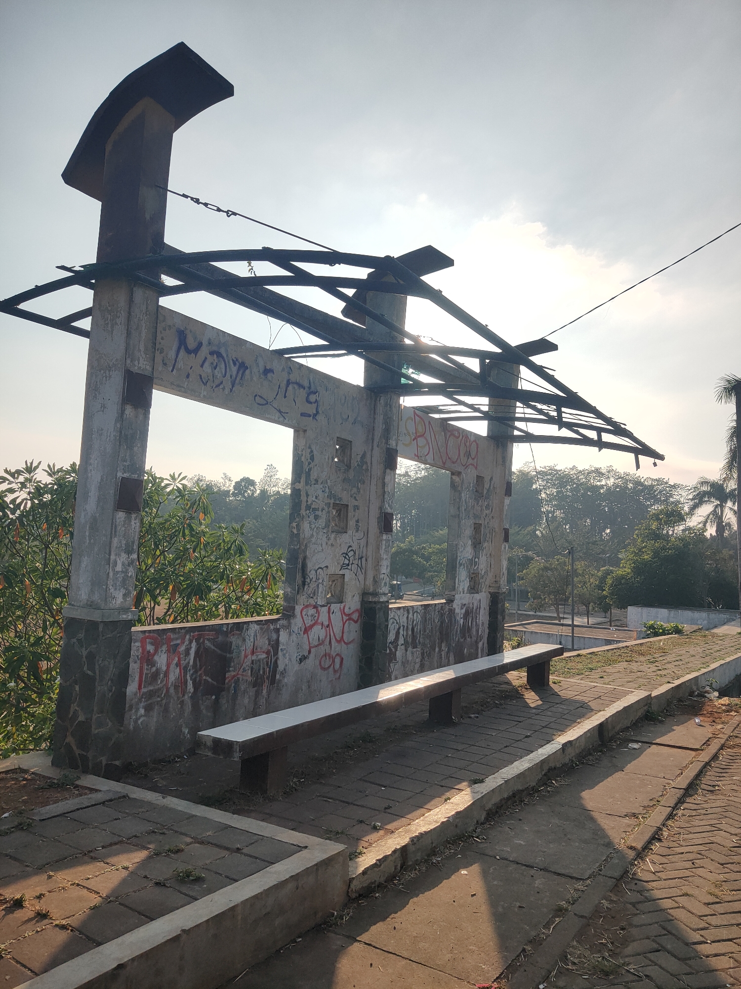 The image shows an outdoor scene with an abandoned, graffiti-covered concrete structure in the foreground. This structure appears to be part of a former shelter or pavilion, with a damaged roof made of metal beams and a concrete bench in front of it. The ground is paved with bricks and concrete. In the background, there is lush green foliage and trees, with a hazy sky suggesting it is either early morning or late afternoon. The location is identified as Kalongan, Indonesia. There are no people visible in the image.