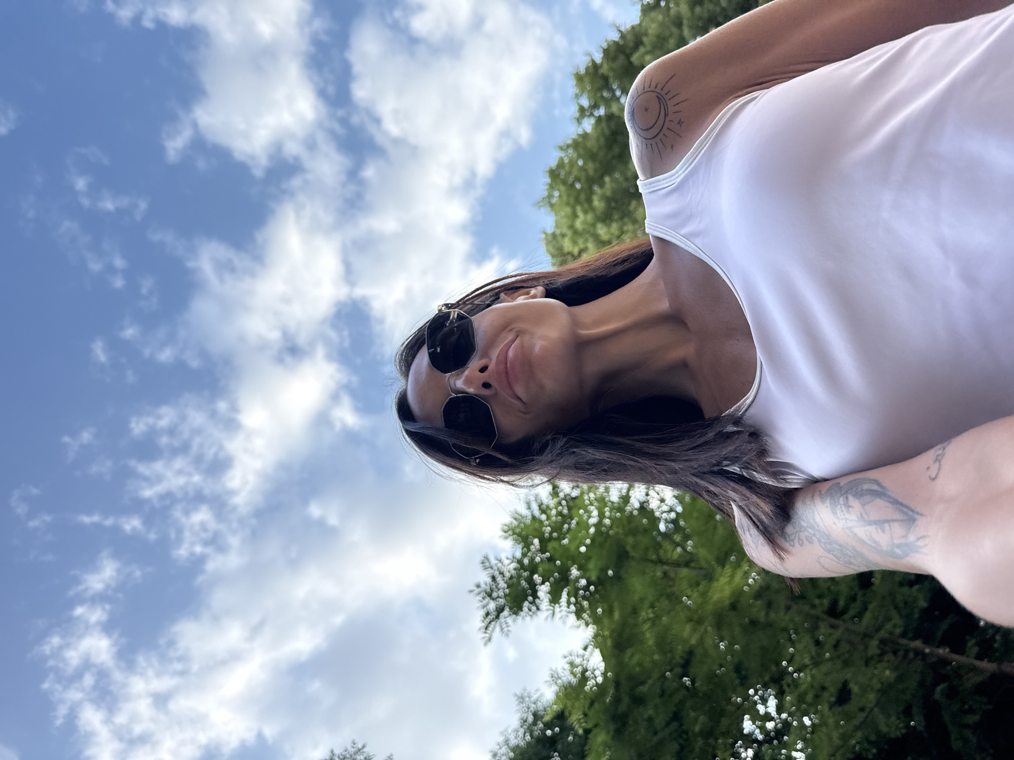 The image is a low-angle, close-up shot of a woman wearing a white tank top and sunglasses, taken outdoors. She has dark hair and visible tattoos on her left arm and right shoulder. Behind her, the sky is blue with scattered white clouds, and green trees are visible. The scene appears to be a casual, outdoor setting. There is no specific information about the location being in the city of Pleven, Bulgaria.