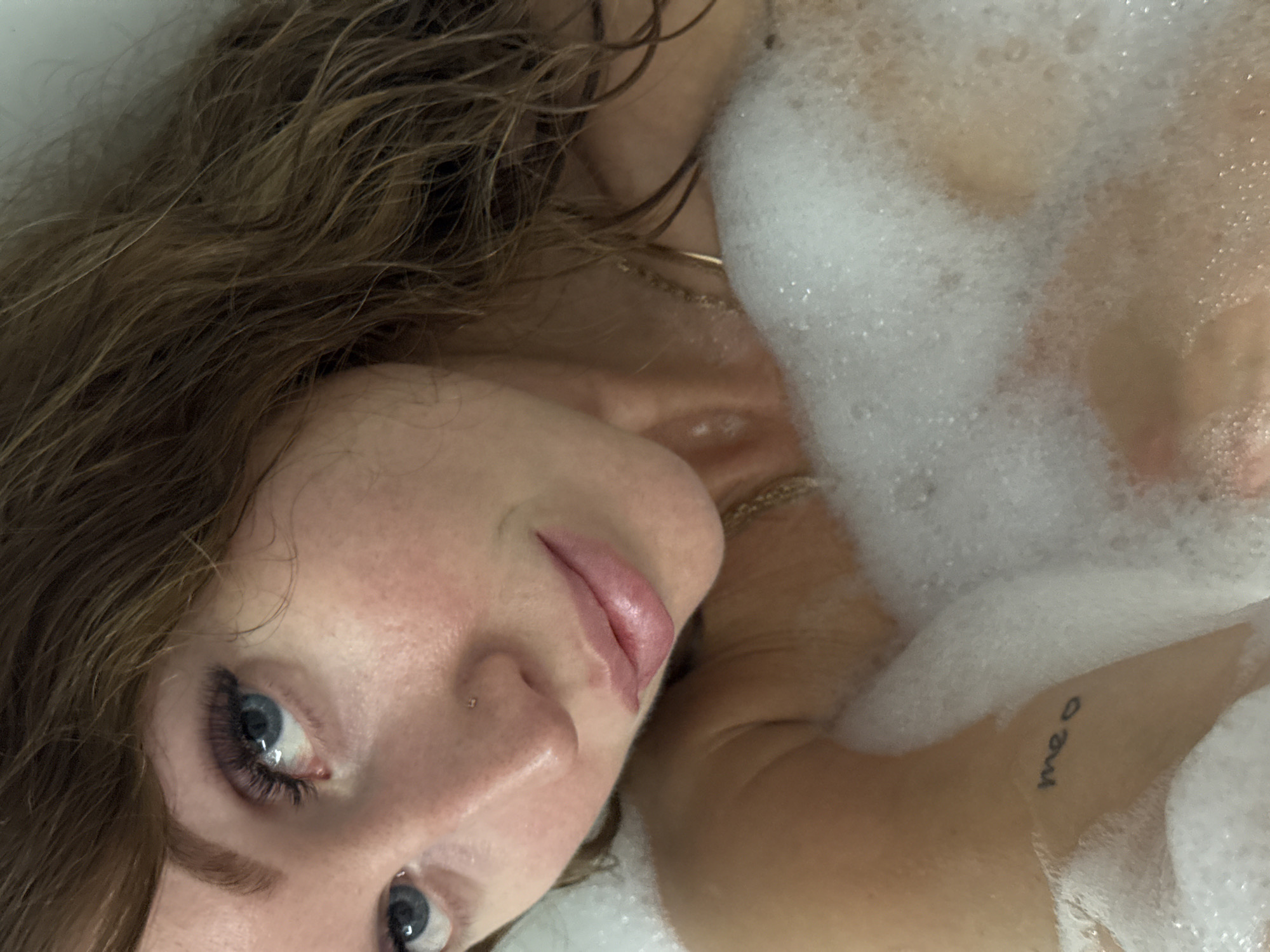 An overhead close-up image shows a person taking a bubble bath in Chaikovsky, Russia. The individual, likely female, is partially submerged, with their head and upper torso visible, largely covered by a thick layer of white foam. Their head is tilted to the left, and they are looking directly at the camera with a calm or neutral expression.
Notable features include prominent blue eyes with long, dark eyelashes, a small stud piercing on the left nostril, and light pink lips. Wet, wavy, reddish-brown hair frames the face and spills around the shoulders. A thin gold chain is visible around the neck. On the upper arm or shoulder, peeking through the bubbles, is a small black handwritten tattoo that reads "meo." The skin visible through the foam appears smooth and lightly flushed. The lighting is soft and even, suggesting an indoor setting.