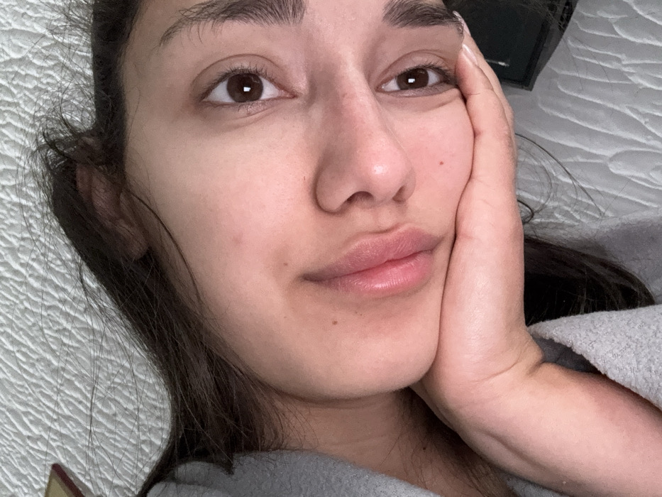 An individual, identified as an adult female, is depicted in a close-up, head-and-shoulders portrait, angled from a slightly overhead perspective. The subject is lying down or reclining, with her head tilted to the viewer's left. Her right hand supports her chin and lower jaw, with the palm making contact with the skin. Her gaze is directed towards the camera. She has dark brown hair, dark brown eyes with visible pupils and catchlights, and full lips that are slightly parted. Her complexion is natural, exhibiting minor moles or blemishes on the cheek. She is wearing a light gray, textured garment, visible on her left shoulder and arm. The background consists predominantly of a light-colored, quilted or textured fabric, consistent with bedding or upholstery. Partially visible in the top left corner is a dark object with a gold-colored edge, possibly a book. In the bottom right corner, a dark, rectangular object is partially obscured by the subject's hand and arm. The lighting is soft and frontal. The image itself provides no specific visual indicators to determine the location in Zankovići, Montenegro.