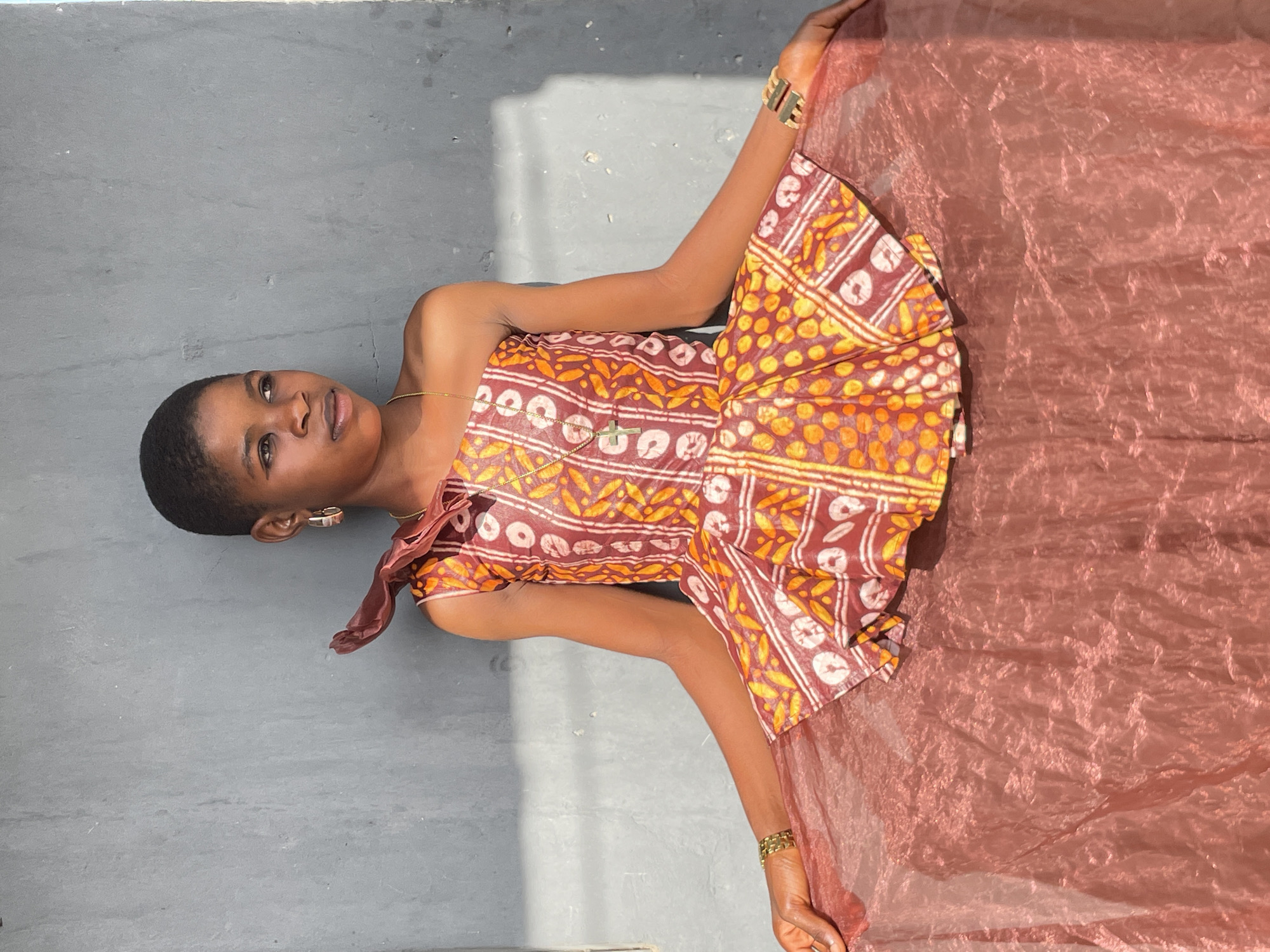 A young person, likely female, with dark skin and short, dark hair, is depicted lying on a gray concrete or paved surface in bright daylight. She is looking upwards towards the camera with a calm and serene expression, a slight smile on her face.
She is wearing a vibrant, short-sleeved dress with a rich brown base patterned with yellow and white or cream geometric and abstract designs. The dress features subtle ruffles or decorative elements around the shoulders and neckline. Her left wrist is adorned with a gold-colored cuff bracelet, and a delicate gold chain with a small cross pendant is visible around her neck. Small earrings are also visible.
Her arms are raised, and her hands are holding a large piece of sheer, shimmery fabric in a reddish-brown or bronze color, which is spread out to her left. The fabric appears translucent and reflects the bright sunlight, creating a luminous effect.
The setting is outdoors in Badagry, Nigeria, under strong natural sunlight, which casts distinct shadows on the gray ground and a nearby wall, suggesting a bright and clear day. The background is simple and uncluttered, focusing attention on the subject. There is no visible text in the image.