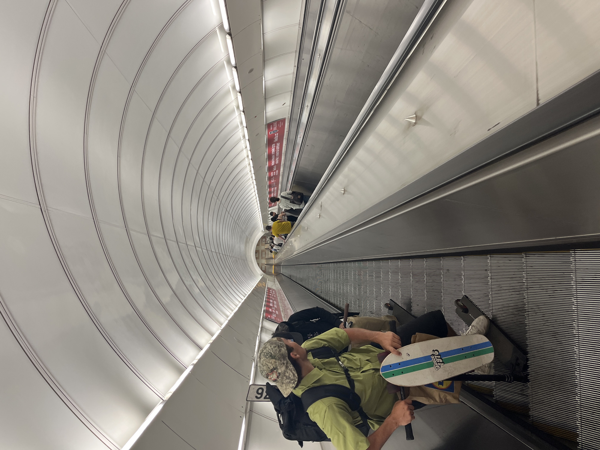 A man is lying down on an escalator inside a metro station in Prague, Czechia. He is wearing a green shirt, khaki pants, a camouflage hat, and has a black backpack. He has a skateboard in his hands. He is surrounded by luggage. There are other people standing on the escalator further up. The escalator is inside a white, rounded tunnel. A sign with the number "92" is visible near the man.