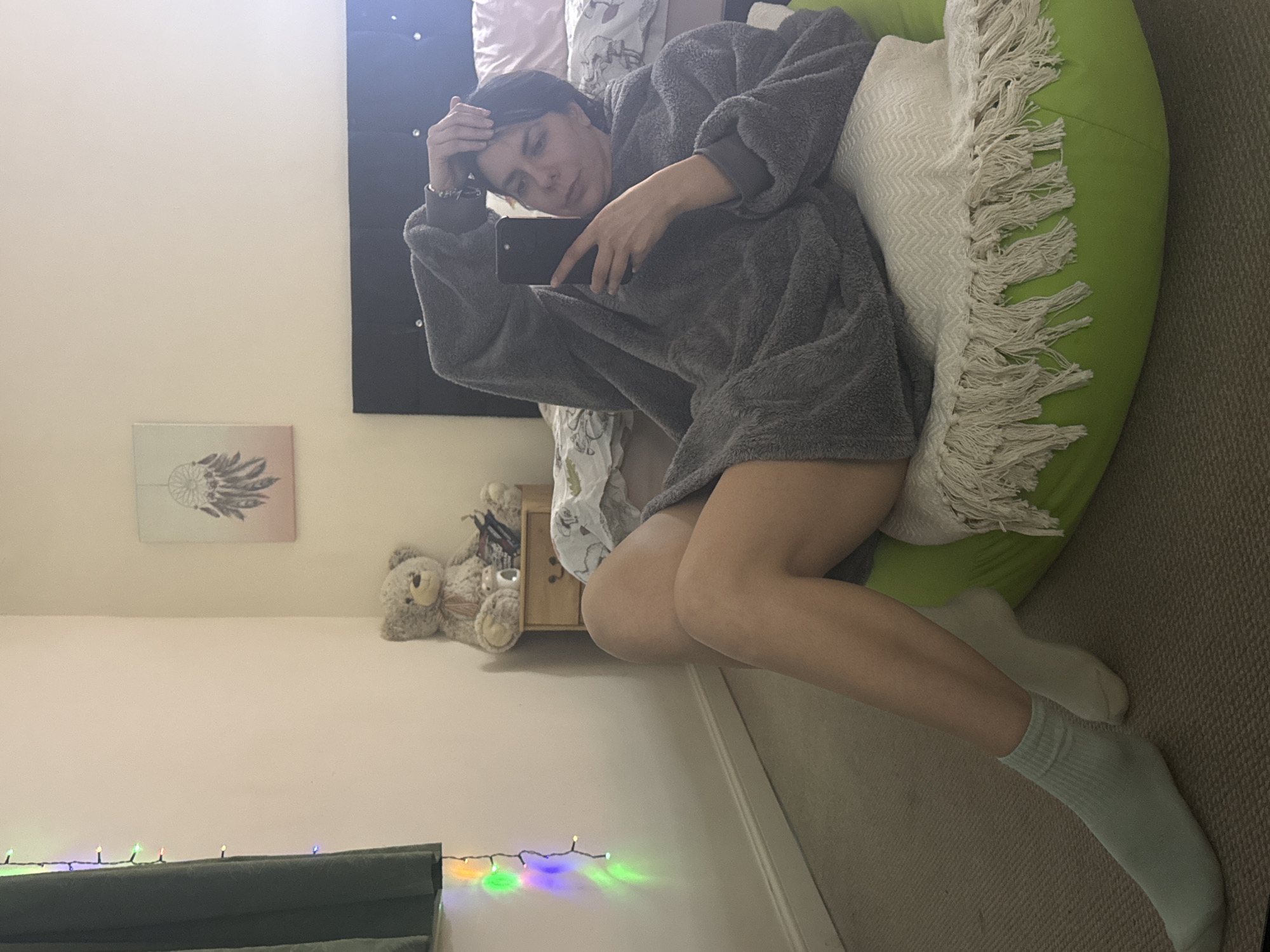 A woman in a gray, oversized sweater and socks is lying on a lime green beanbag chair in a bedroom. She is holding a smartphone in her left hand and her right hand is touching her head. The room contains a bed with a black headboard, a bedside table, a teddy bear, and a wall decoration featuring a dreamcatcher design. String lights are visible along the bottom of the image. The image does not provide any information to determine the location in the city of Great Yarmouth, United Kingdom.