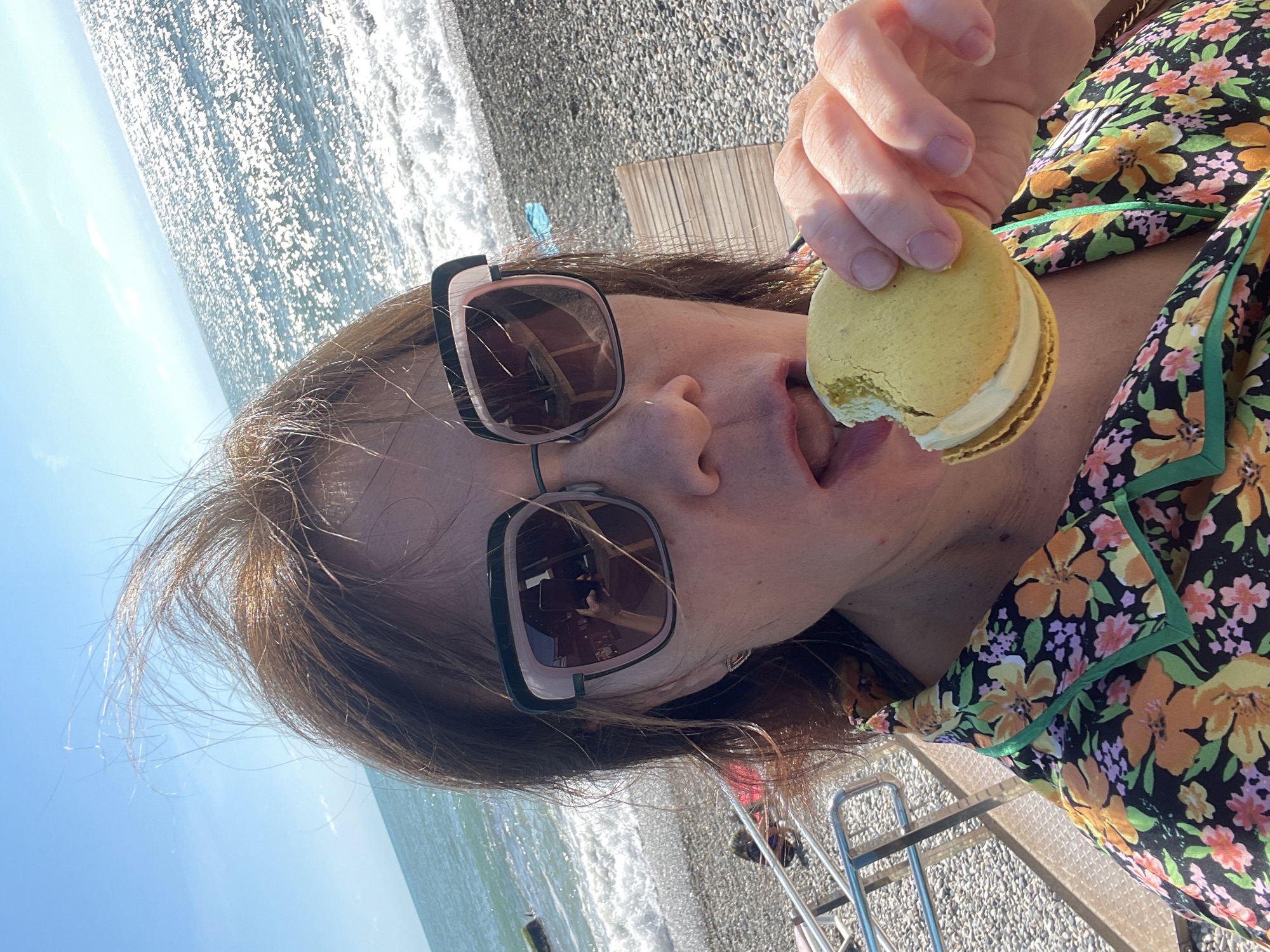 A woman with brown hair is taking a selfie on a beach, eating a yellow macaron. She is wearing large, square sunglasses and a black shirt with a floral pattern featuring orange and pink flowers and green trim. The background shows the sea and the sky. In the reflection of her sunglasses, another phone can be seen being held to take the picture. The beach is made of pebbles, and a wooden deck chair is visible behind her. The woman appears to be at a beach resort in Sochi, Russia, given the clear sky and sunny weather typical of the region.