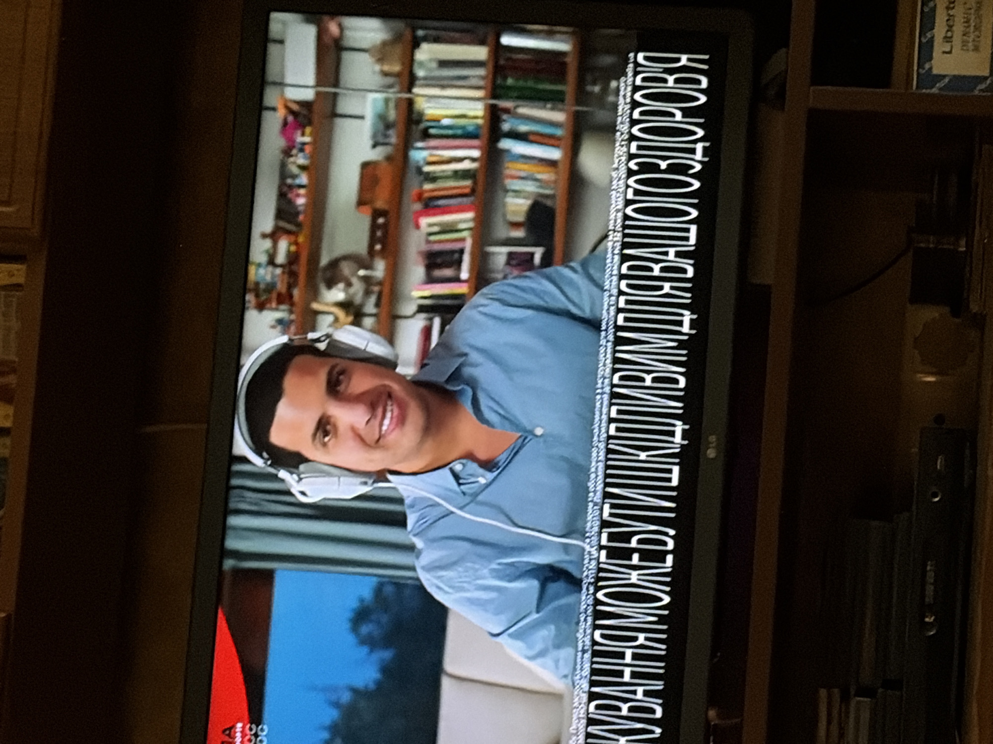 A man wearing headphones is seen from the chest up, smiling and looking at the camera. He is wearing a blue button-down shirt. He is sitting in front of a bookshelf filled with books. A caption in white text with a black outline appears at the bottom of the image, featuring Cyrillic lettering. The background behind him appears to be a room with a couch and a window with blue curtains. The scene is captured on a television screen, with the lower part of the television and some furniture visible below the screen.