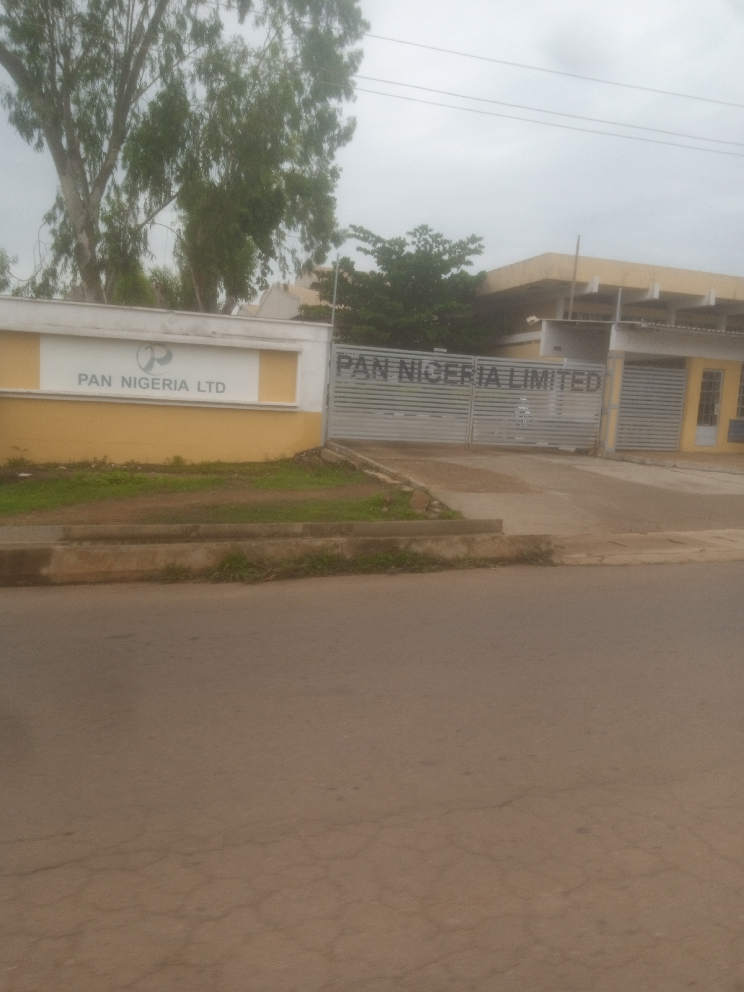 The image shows the front entrance of a facility labeled "PAN NIGERIA LIMITED" in Kakuri Gwari, Nigeria. The entrance features a large metal gate with horizontal slats. To the left of the gate is a white wall with a yellow section beneath it, displaying the "PAN NIGERIA LTD" logo and text. Behind the fence, a building with a pale yellow roof and white pillars is visible. Tall trees with green foliage are present on the left side of the image, in front of the building. The ground in front of the facility is a dirt pathway with some sparse grass. A paved road occupies the foreground of the image. The sky is overcast.