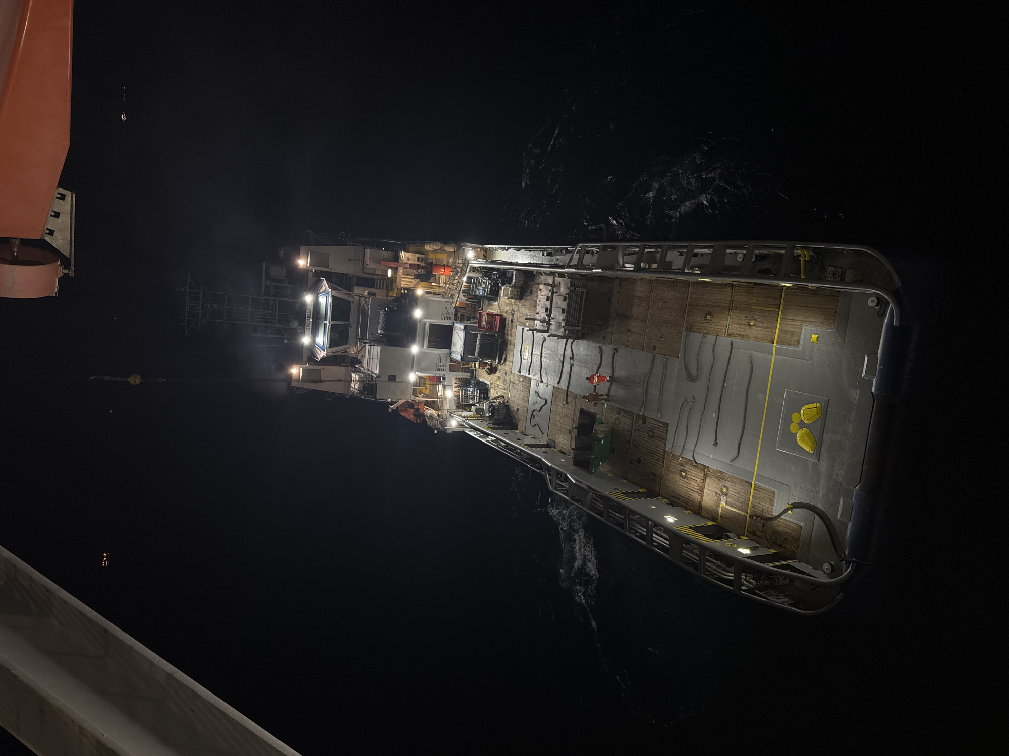 This image, captured at night, shows an aerial view of a large vessel with its deck illuminated. The vessel, identified as the "IEVOLA BLUE" by lettering on its bridge, is positioned in dark water, with some minor waves visible. The deck of the ship is a mix of wooden and metal surfaces, with various equipment and materials stowed. Two yellow, boat-shaped buoys are present on the deck.
Two figures in orange safety suits are visible on the deck. One is standing near the stern, and the other is closer to the center of the vessel. Several vehicles, including what appears to be a red truck and other machinery, are also on the deck. The bridge of the ship is brightly lit, with windows offering a view into the interior.
In the background, the night sky is dark, with a few distant lights visible on the water, possibly from other vessels or structures. Part of a large orange crane structure is visible in the upper right corner of the image, and a white structural element extends from the upper left corner, suggesting the photograph was taken from a higher vantage point, possibly another vessel or a crane. The overall scene depicts activity on a ship at sea during nighttime operations. The location is not specified in the city of Unknown, Italy.