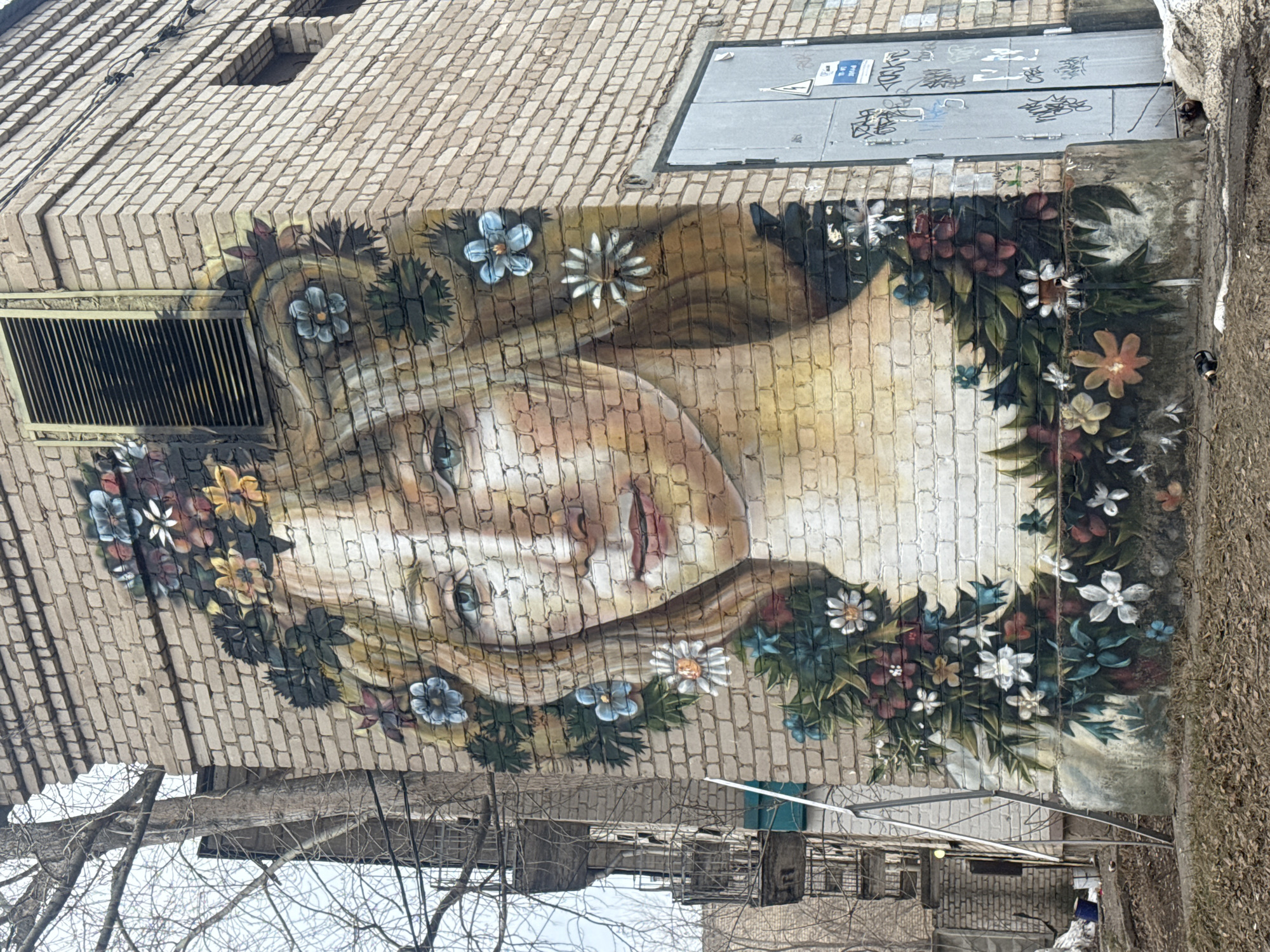 A vertical eye-level medium shot shows a mural of a woman wearing a wreath of flowers painted on a brick building in Chaikovsky, Russia. The woman is facing down and to the left with a neutral expression. Her eyes are blue. Her hair is long and blonde. The wreath of flowers is made of various colors, including white, blue, yellow, and red. The building is made of light brown bricks. There is a vent above the woman's head and a sealed door to the right. Trees are visible at the bottom of the frame. The ground is covered in brown leaves. The lighting is diffuse.