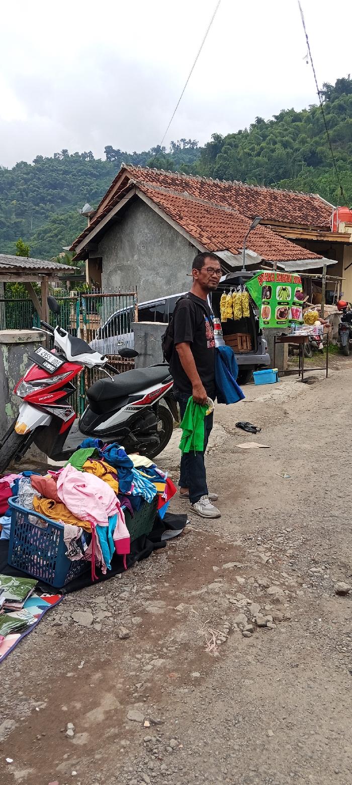 The image shows a man standing in front of a small shop in Kutawaringin, Indonesia. He is wearing a black t-shirt and blue jeans. He is holding a green bag in his right hand and a blue scarf in his left hand. He is looking at the camera. The shop has a sign in Indonesian that says "Fried Banana". There is a motorcycle parked in front of the shop. The motorcycle is red and white. There is a pile of clothing and a blue basket on the ground in front of the motorcycle. There is a pile of bananas on the table. There are green hills in the background. The scene is located at -6.974 degrees latitude and 107.519 degrees longitude.