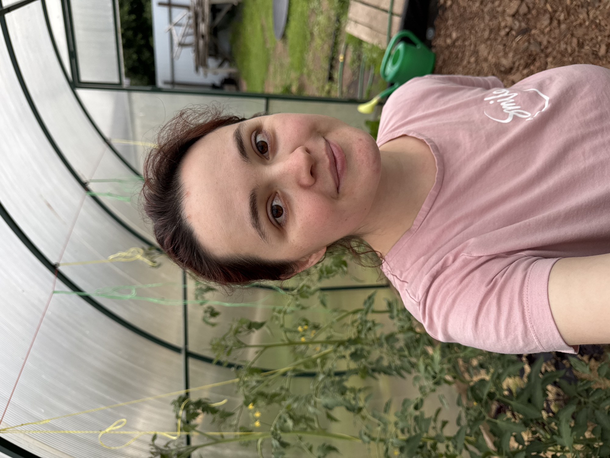 A woman with shoulder-length brown hair and brown eyes is taking a selfie in a greenhouse. She is wearing a pink t-shirt with the word "Smile" printed in white on it. Behind her, tomato plants are growing, with some small yellow flowers visible. A green watering can is visible in the background, along with some wooden planks and a grassy area. The greenhouse has a clear plastic roof and green metal supports. The lighting suggests it is daytime. The woman is looking at the camera with a slight smile.