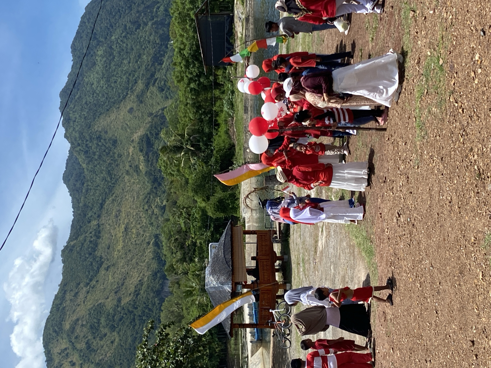 The image shows a group of people, many of whom are wearing red and white attire, gathered outdoors near a body of water. In the background, lush green mountains rise under a bright blue sky with scattered clouds. A small, traditional-style building with a thatched roof is situated on the shore of the water. Several flags are visible, including a large yellow and white flag and smaller flags with red and white stripes. Red and white balloons are also being held by some individuals. The ground is covered in gravel and some patches of grass. In the foreground, a young child in a red outfit is walking away from the camera. The scene appears to be a festive gathering or celebration in a natural, scenic setting. The location is identified as Aneuk Paya, Indonesia.