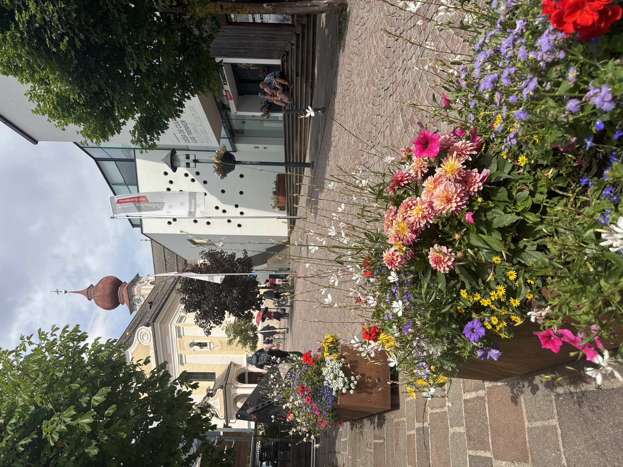 The image depicts a street scene in Ortisei, Val Gardena, Italy. In the foreground, large planters overflow with a vibrant assortment of flowers, including pink and orange dahlias, purple petunias, yellow daisies, and delicate white blossoms. The planters are made of a dark, rustic metal. The ground is paved with cobblestones.
In the mid-ground, a street extends into the distance, lined with buildings. On the left, a yellow and pink church with a red onion dome and clock tower is visible, partially obscured by trees. Adjacent to the church is a modern white building with a glass facade and a prominent flag bearing the text "Dolomites Val Gardena". Another building to the right is also modern, with a large glass entrance and stairs leading up to it. On these stairs, two individuals, a man and a woman, are seated facing each other.
In the background, several people are walking along the street. One person is riding a bicycle. A red umbrella is held by someone in the crowd. The sky is partly cloudy. The overall impression is of a pleasant, well-maintained town square on a sunny day.