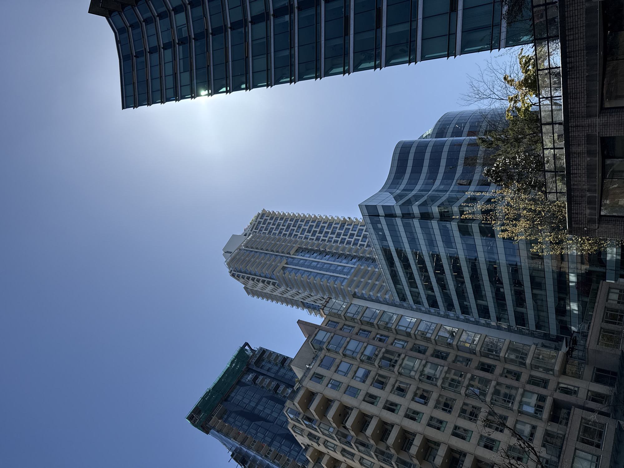 The image presents an upward-looking perspective of multiple tall, modern buildings against a bright, clear blue sky in what appears to be Vancouver, Canada. The dominant subjects are the skyscrapers, showcasing a variety of contemporary architectural styles.
At the top of the frame, a sleek, dark-glass building with prominent horizontal elements is visible, its facade reflecting the sun's glare. To its right, another distinctive building features a wavy, curvilinear glass facade, creating a dynamic, fluid appearance. Further down and to the left, a very tall residential-style tower with numerous balconies is visible, characterized by its lighter-toned and structured design. In the lower left quadrant, a building under construction or renovation is partially seen, covered with green safety netting. Adjacent to it, towards the lower right, an older-style building with a grid of square windows and concrete elements adds contrast to the more modern structures.
The sky is a vibrant, cloudless blue, indicating clear weather. A strong sunburst is visible towards the upper left, suggesting bright daylight, likely midday or early afternoon. This intense light creates strong reflections on the glass surfaces of the buildings, mirroring the sky and hints of surrounding urban elements. Sparse tree branches and some green foliage are visible, either growing between buildings or reflected in their glass panels. There is no specific activity or event taking place; the image captures a static architectural landscape from a worm's-eye view, emphasizing the verticality and scale of the urban environment. No visible text is present in the image.