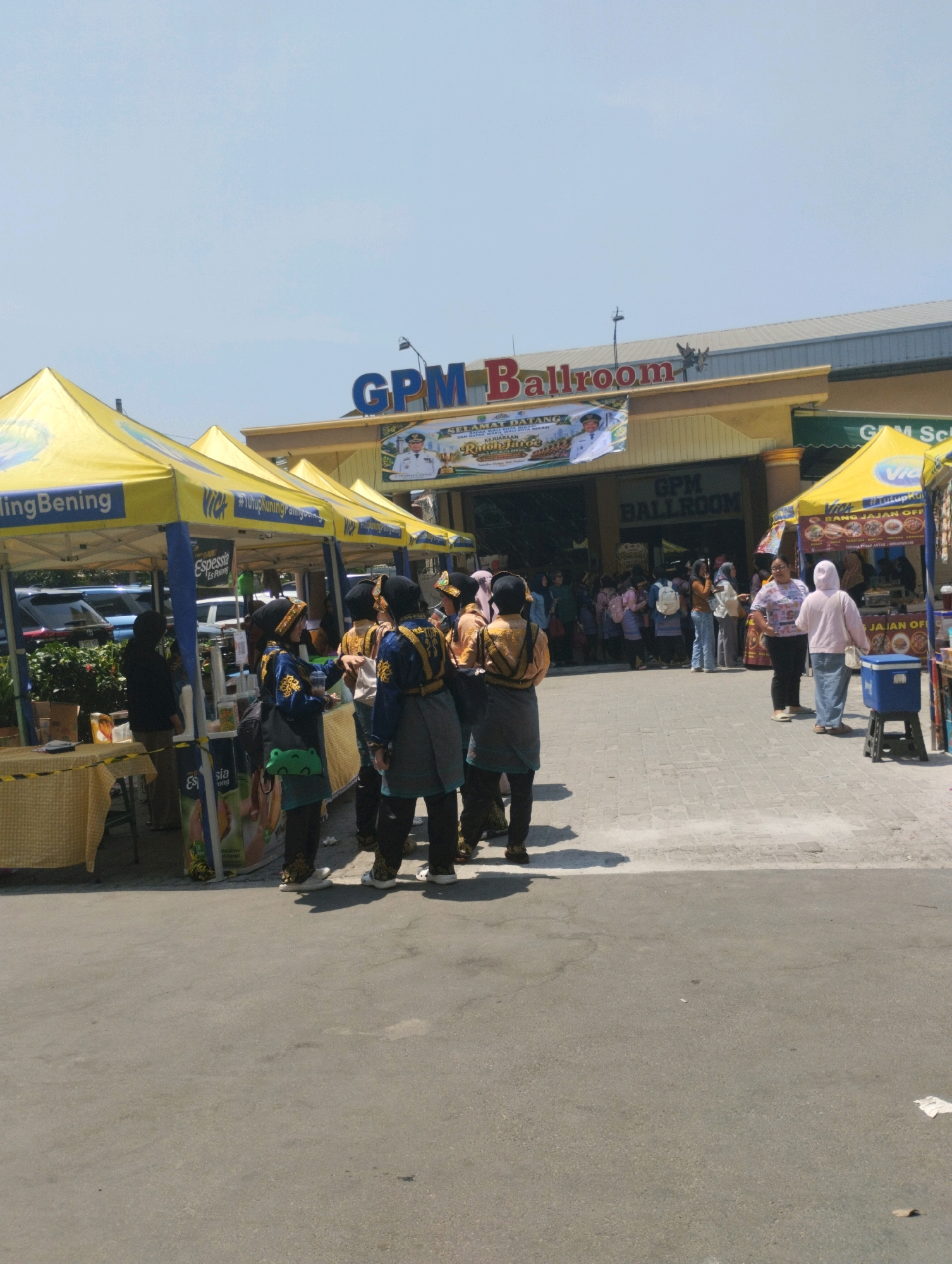 The image shows the exterior of the GPM Ballroom in Bekasi, Indonesia, on a clear day. A crowd of people is gathered in front of the building, with several vendors set up outside under yellow tents. Some individuals are wearing traditional Indonesian attire. One tent displays banners advertising "Vick" and a food stall. The overall scene suggests an event or market is taking place.