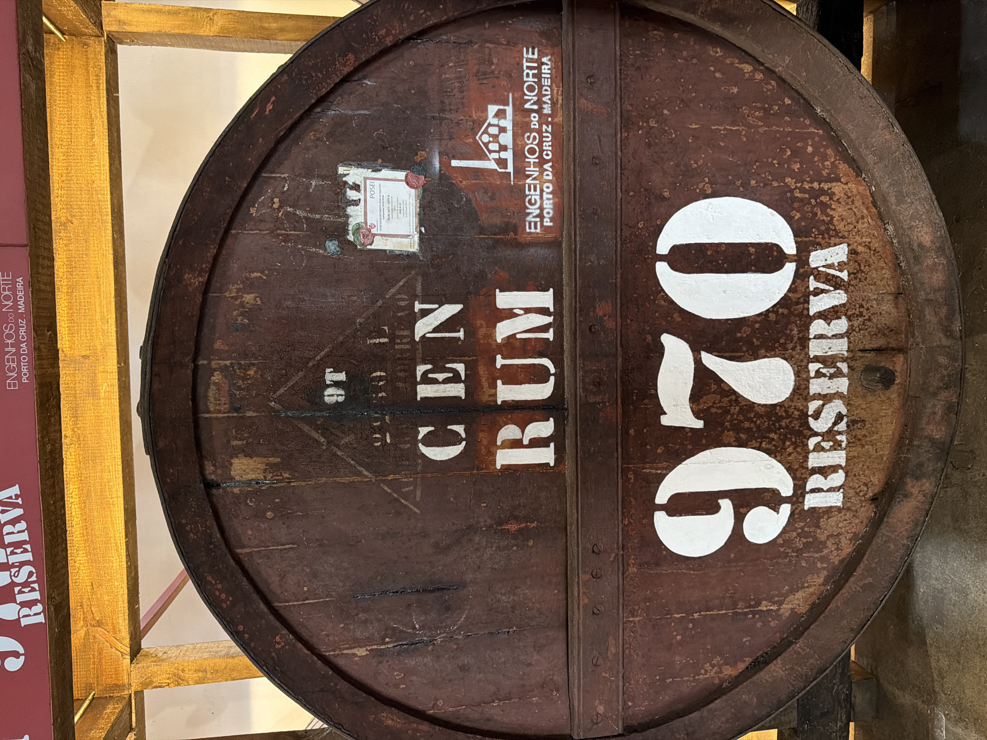 A medium shot depicts a large wooden barrel with prominent white lettering and markings. The words "CEN RUM" are stacked vertically in the center. Above them, a smaller logo with the text "ENGENHOS DO NORTE" and "PORTO DA CRUZ. MADEIRA" (the city of Porto da Cruz is within the Machico municipality of Portugal) can be seen along with what appears to be a small information label. To the right, the large number "970" is vertically aligned alongside the word "RESERVA". The barrel has metal bands and rivets holding it together, and the wood shows signs of aging and wear. The barrel is likely used for storing or aging rum. Part of what appears to be a wooden frame and the corner of a display with the word "RESERVA" is visible on the left side of the frame.
