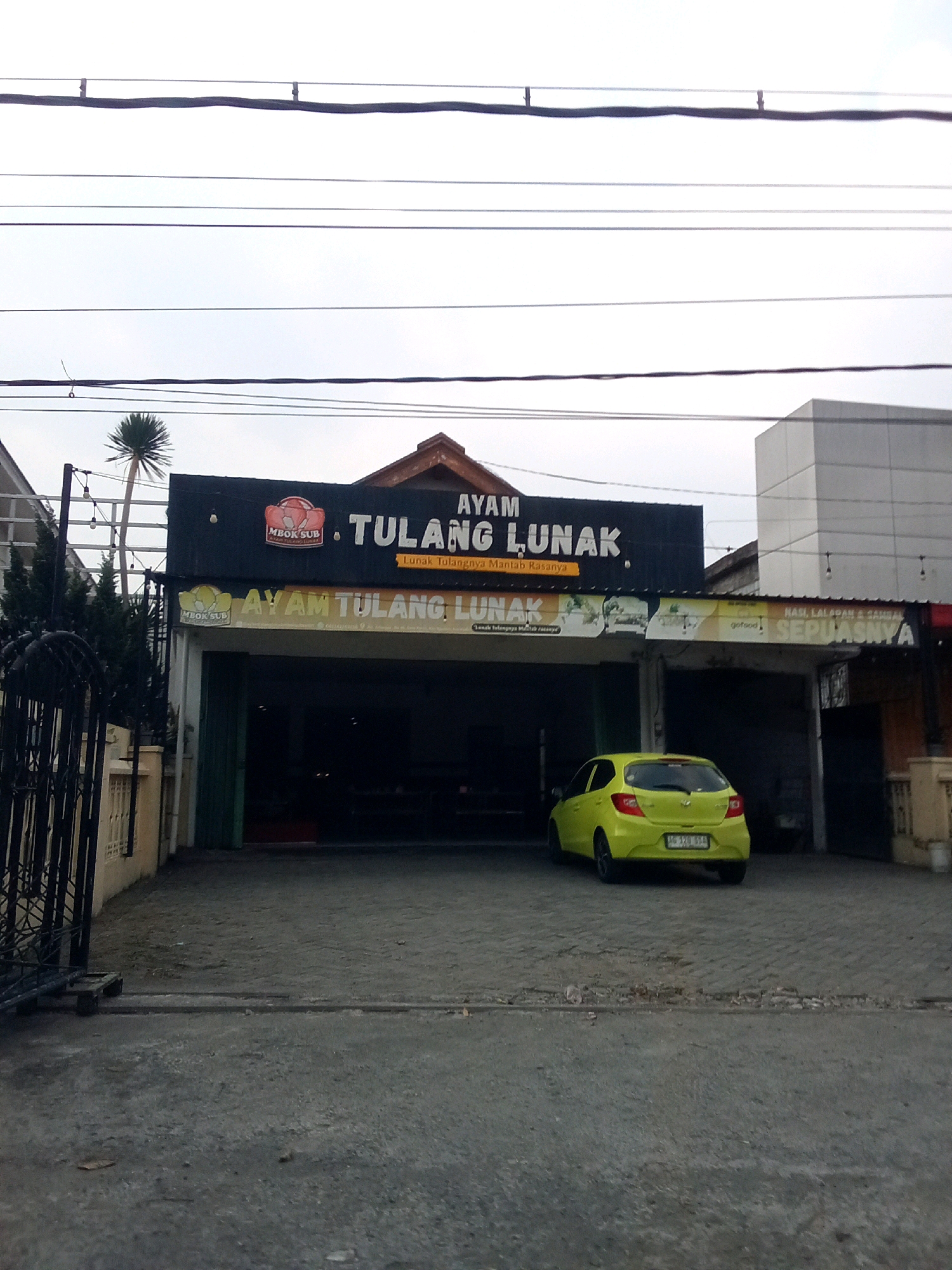 The image shows a storefront in Ngasem, Indonesia, with a prominent sign that reads "AYAM TULANG LUNAK". Below the main sign, a smaller banner states "Lunak Tulangnya Mantap Rasanya". A bright yellow car is parked in front of the building. The sky is overcast. The building appears to be a restaurant.