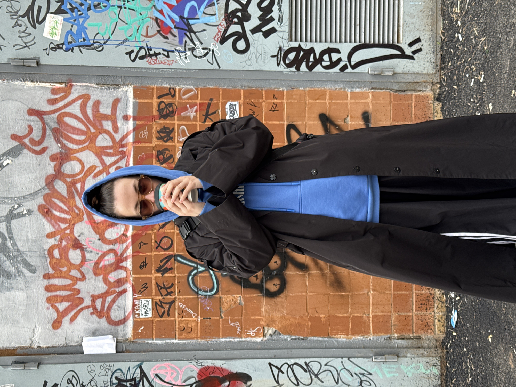 A person wearing a blue hooded sweatshirt and a black trench coat is holding a cup and standing in front of a wall covered in graffiti. The wall is composed of different sections, with graffiti in various colors and styles. The bottom section of the wall is brick-like with some graffiti. The location appears to be an urban area based on the presence of graffiti. The city and country are Chaikovsky, Russia.