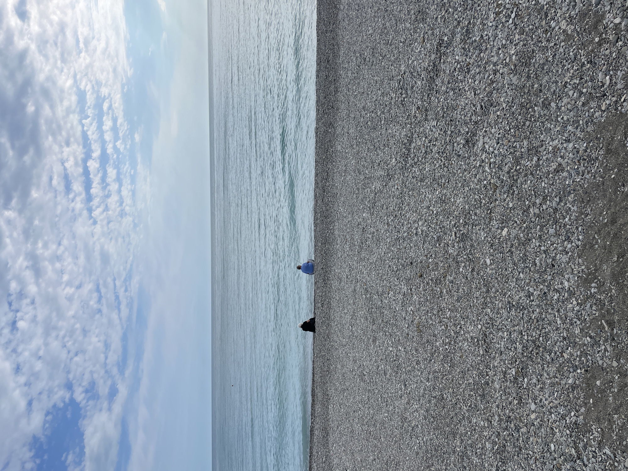 The photograph depicts a coastal scene in Olginka, Russia. The image is vertically oriented. A cloudy blue sky occupies the upper left corner. The sea stretches across the central portion of the frame. Two figures are present near the water's edge. One is wearing a light blue shirt and is standing. The other is wearing dark clothing and is crouching. A pebble beach fills the right side of the image.