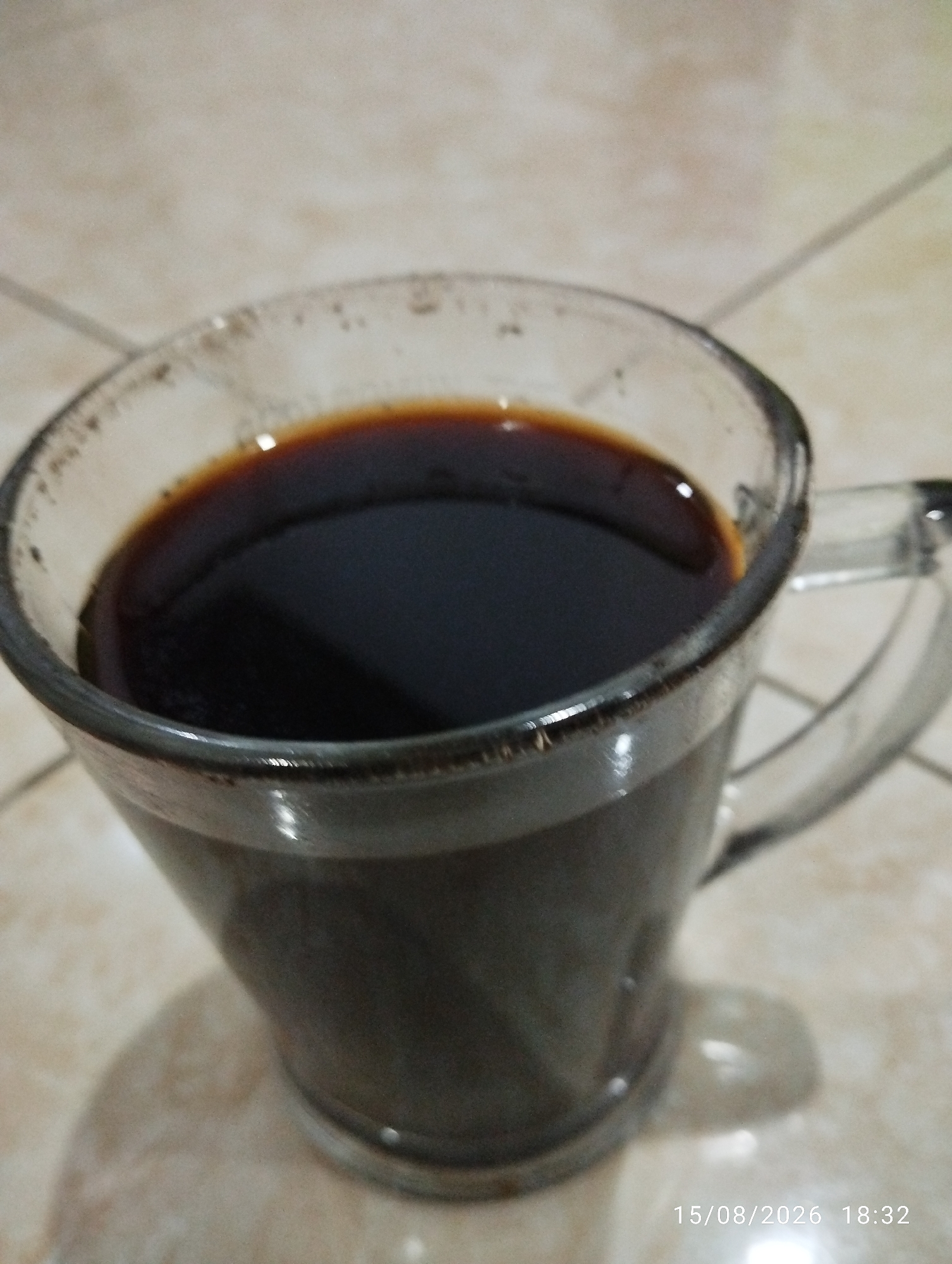 A clear glass mug filled with dark liquid, likely coffee, sits on a tiled surface. The mug has a handle and is slightly angled towards the viewer. The liquid surface is smooth and reflective, with a thin rim of darker color at the top. The background consists of light-colored, square tiles with visible grout lines. A shadow of the mug is cast on the tiled surface. A timestamp "15/08/2026 18:32" is present in the bottom right corner of the image. There are no people or animals present in the image. The scene is indoors, based on the lighting and the tiled floor. The location is specified as Sukamaju, Indonesia.