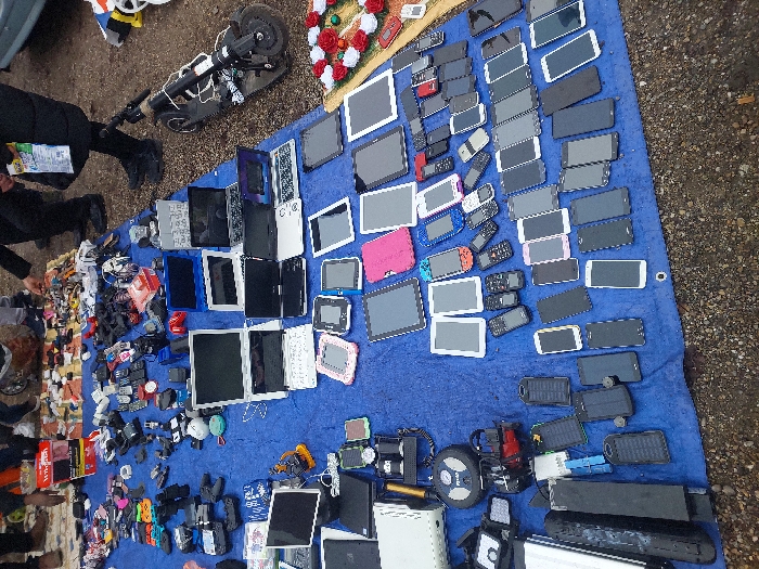The image depicts an outdoor market stall in Eckbolsheim, France, showcasing a large array of used electronic devices meticulously arranged on a royal blue tarp laid over a gravel ground surface. The upper portion of the tarp features various laptops, both open and closed, alongside smaller electronics such as gaming controllers, smartwatches, and assorted chargers. The mid-section displays numerous tablets, some enclosed in colorful protective cases. The lower two-thirds of the tarp are densely covered with multiple horizontal rows of mobile phones, encompassing a wide range of smartphones in various models and colors, interspersed with older feature phones. Other visible objects include several black solar power banks, a portable air pump, and a white game console unit. In the upper right quadrant, a black electric scooter rests on its side adjacent to a small arrangement of artificial red and white roses. Several partially visible individuals are present in the background, standing near the stall, with one person's legs, lower torso, and arm distinctly visible near the scooter and the displayed items.