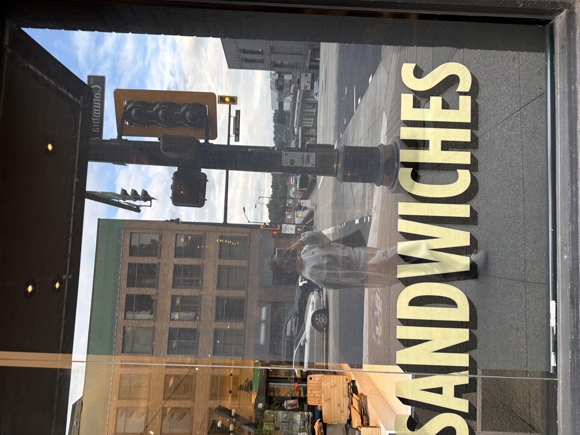The image shows a reflection in a storefront window in New Westminster, Canada, with the word "SANDWICHES" in large gold lettering across the bottom. Through the window, a brick building with multiple stories and windows is visible, as is a traffic light and street sign for "Columbia." A person is reflected in the window, holding a smartphone to take a picture. Several cars and a person walking on the sidewalk are also visible in the reflection of the street. The sky above is cloudy.