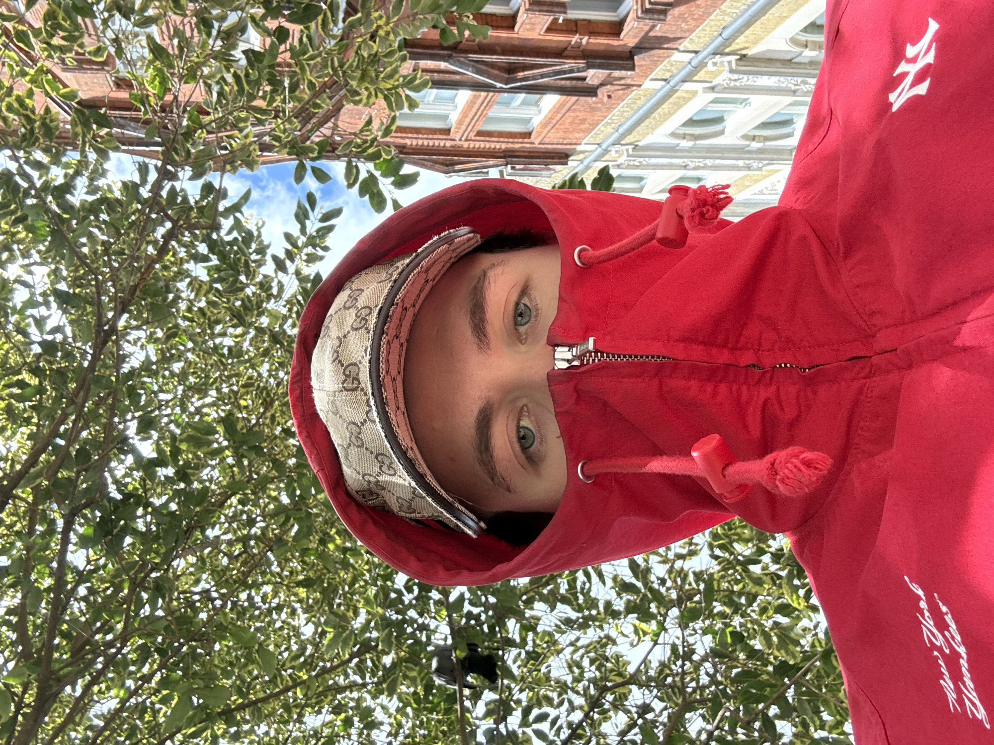 This media file captures a unique, low-angle portrait of an individual outdoors in London, United Kingdom. The subject's face is seen looking directly upwards into the camera, with only their eyes, eyebrows, and forehead clearly visible, revealing light-colored eyes. They are wearing a bright red hooded sweatshirt, which is zipped up, and a beige baseball cap with a dark brown monogram pattern, identifiable as the Gucci "GG" logo, is worn beneath the hood. The red sweatshirt also features white embroidered text on the wearer's right side, reading "New York Yankees™".
The background is a vibrant mix of natural and urban elements. Lush green foliage from a tree, with some leaves showing variegated yellow-green patterns, frames the subject densely in the foreground and mid-ground. Through the gaps in the leaves, a clear blue sky with scattered white clouds is visible, indicating a sunny daytime setting. In the upper background, traditional brick buildings with ornate white window frames and architectural details are present, suggesting an established urban environment consistent with London. The unusual perspective provides an intimate and engaging view of the subject against a characteristic city backdrop.