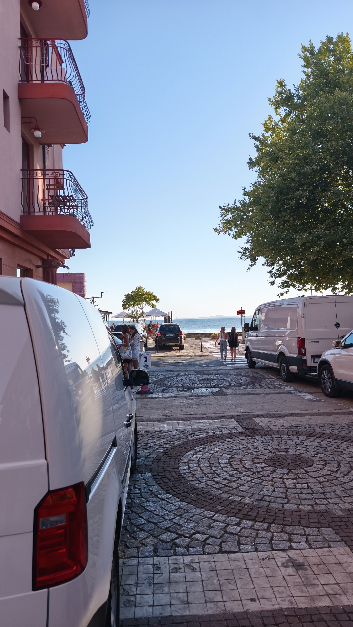 The image depicts a street scene in Pomorie, Bulgaria, with a view towards the sea. In the foreground, the rear of a white van is prominently visible on the left. The street is paved with cobblestones arranged in circular patterns.
On the right side of the street, a white van is parked, and next to it, a white car. Further down the street, a black SUV is parked. In the background, two women are walking across a pedestrian crossing. A few people are seated at tables under umbrellas near the beach. The sea and a clear blue sky form the backdrop.
To the left of the street, a building with multiple balconies, featuring red railings and accents, is visible. A large tree with green foliage is on the right side of the image, partially obscuring the sky. The overall scene suggests a sunny day in a coastal town.
