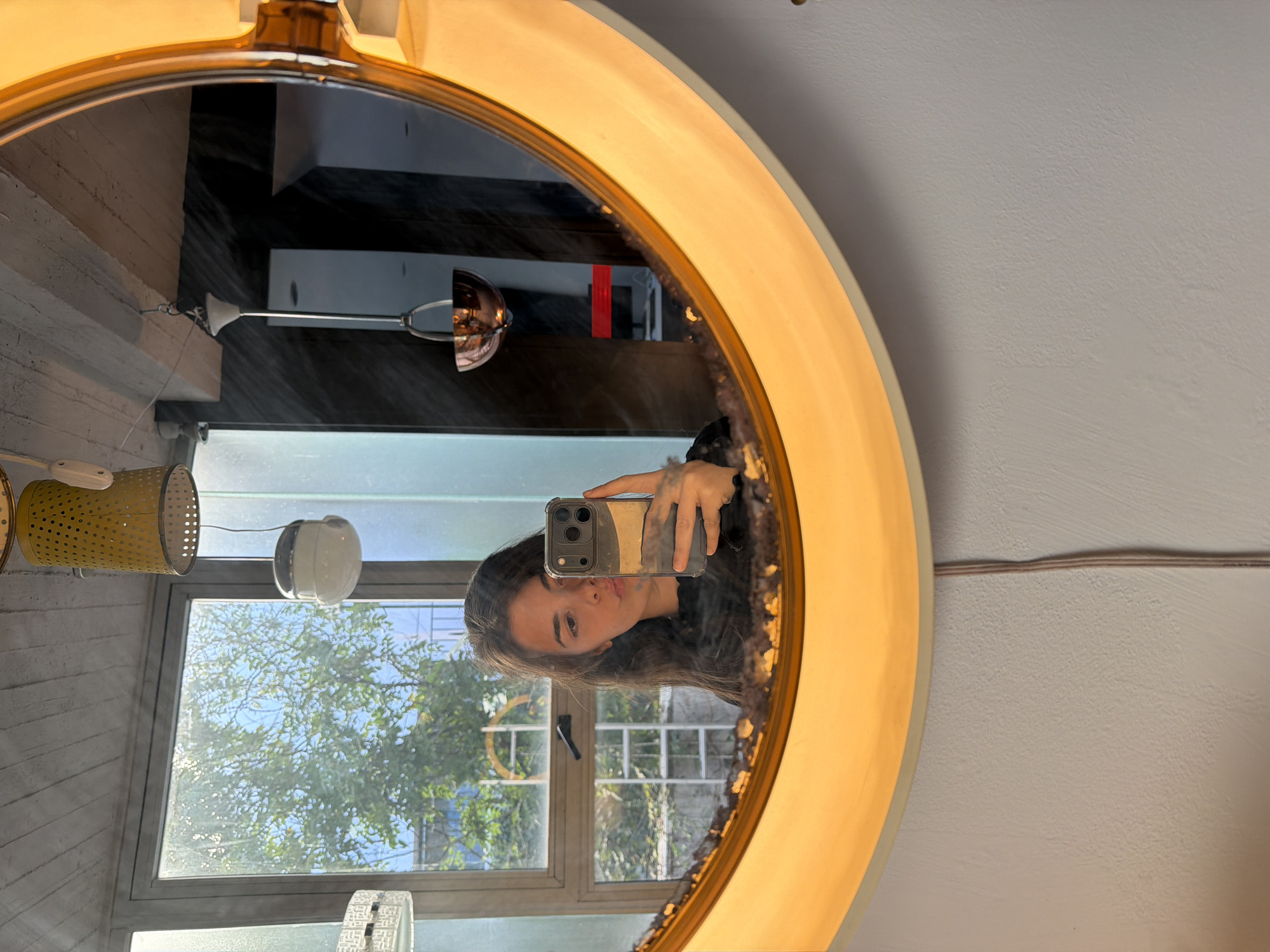 A young woman is captured in a mirror selfie, seen in the reflection of a large, circular mirror with an illuminated golden-yellow frame. The image is rotated 90 degrees counter-clockwise. The woman, with long dark hair, holds a smartphone with a clear case in front of her face to take the picture, her gaze directed towards the camera with a neutral expression.
The interior setting, reflected in the mirror, features textured gray walls, possibly concrete. Various light fixtures are visible, including a perforated yellow cylindrical lamp and a white, dome-shaped light. A window in the background reveals a bright, sunny daytime scene with green trees and a clear blue sky, indicating good weather. Sunlight streams into the room. The mirror's reflective surface shows some smudges, particularly along the lower right edge of the frame. The scene suggests an indoor location, such as a home or a well-lit commercial space, possibly in Buenos Aires, Argentina, as per the provided context. No discernible text is visible in the image.