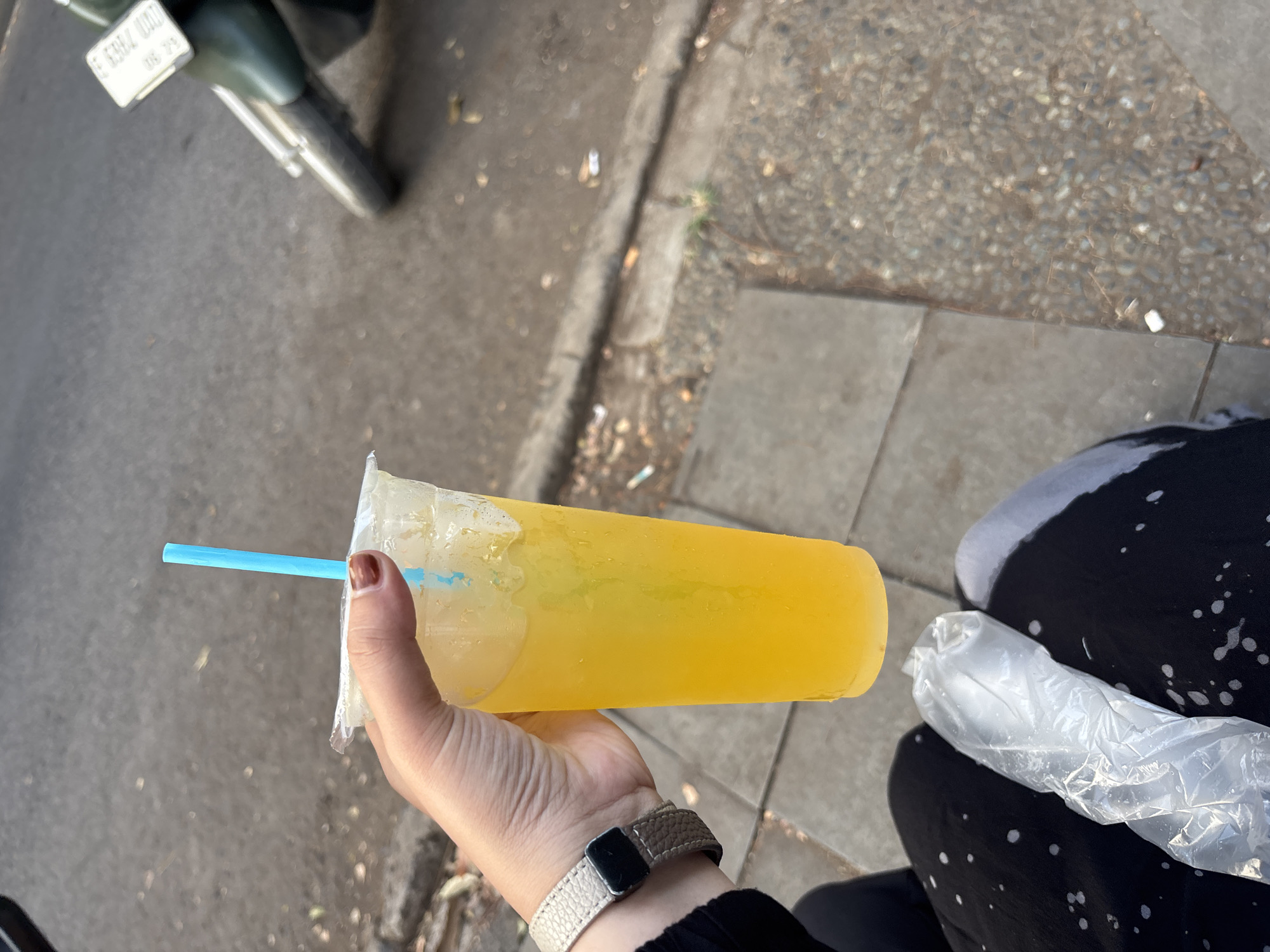 A person's hand, adorned with a brown-polished fingernail and a light-colored wristwatch with a black rectangular face, holds a clear plastic cup filled with a bright yellow beverage. A blue plastic straw extends from the cup. The cup and its contents are held in the foreground, with the background showing a paved walkway and a gray asphalt road. A motorcycle with a white license plate is partially visible on the right side of the image. The overall scene suggests an outdoor setting, possibly on a street in Cirebon, Indonesia, given the presence of a motorcycle commonly used for transportation in the region.
