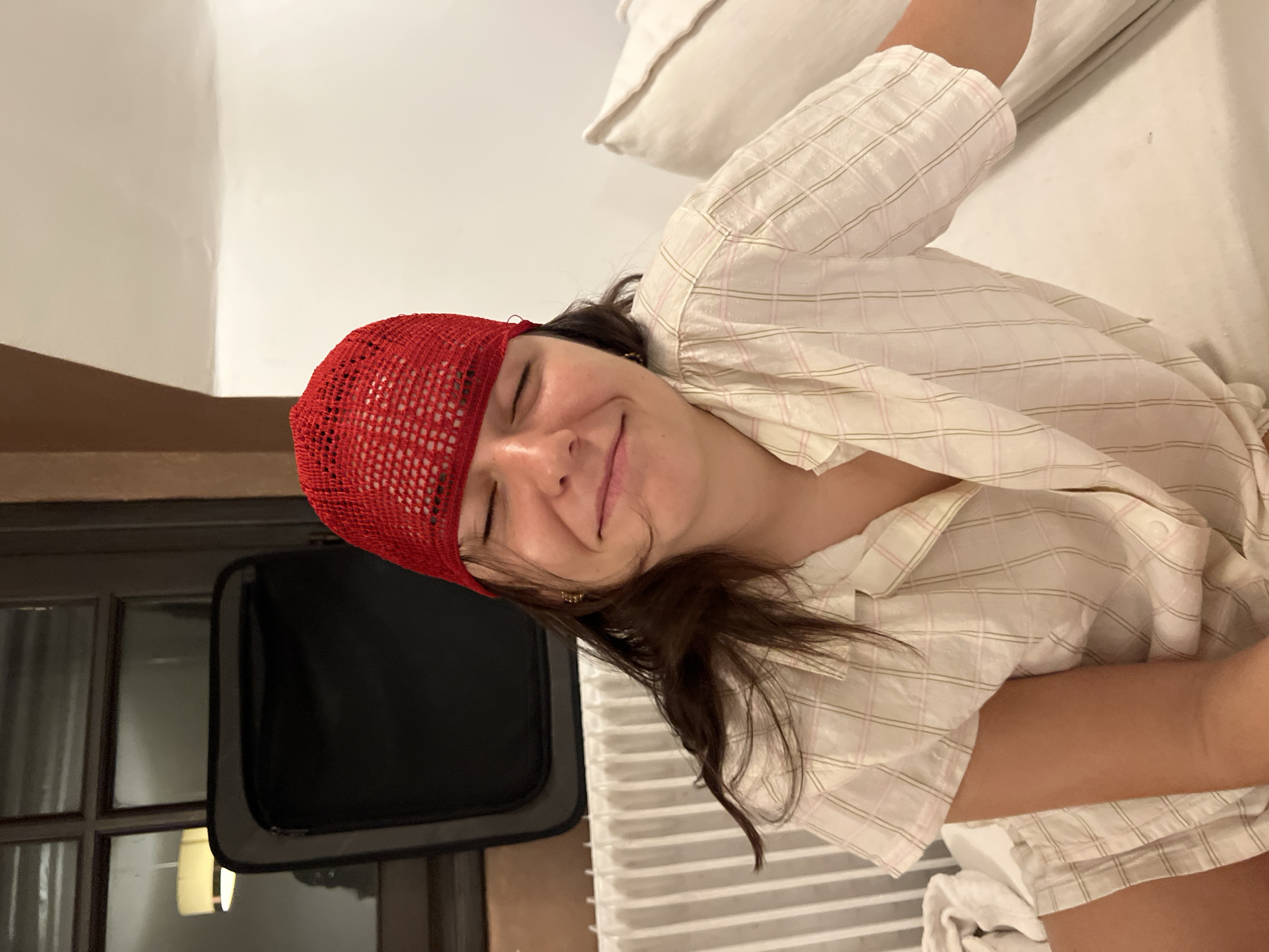 A medium, rotated shot shows a smiling woman with her eyes closed, wearing a red crocheted cap and a plaid, collared shirt. She appears to be lying on a bed with a white pillow behind her. To the left, there's a dark television screen and a wall. It is unlikely to ascertain the location in Paris from the image.