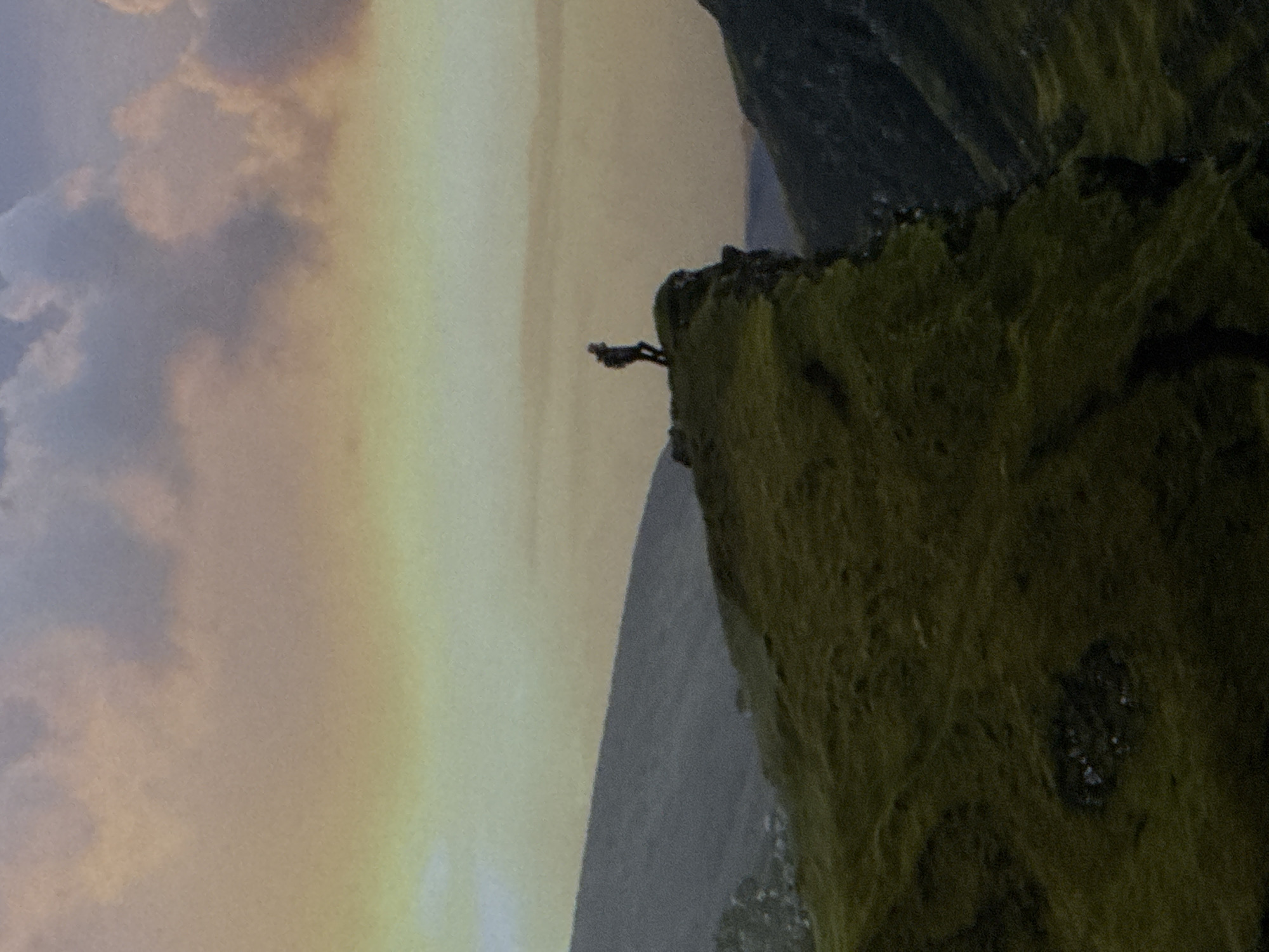 A lone figure stands at the edge of a grassy cliff overlooking a valley. The sky above is a mix of soft peach and pale yellow clouds. The scene is a natural landscape with rolling hills and mountains.